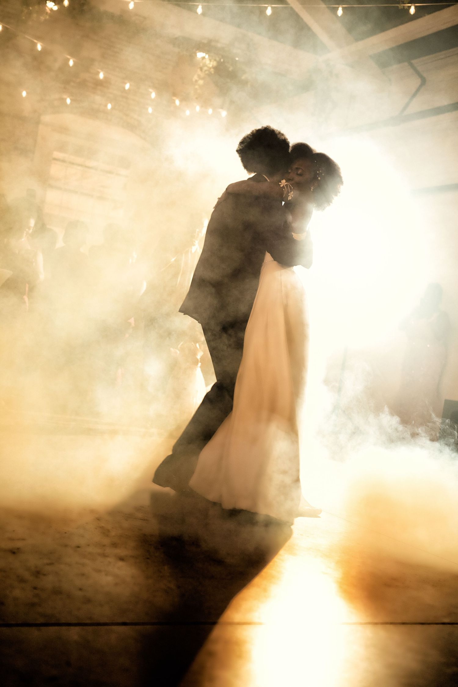 Bride and groom share first dance surrounded by romantic fog and warm golden light in a rustic wedding venue.