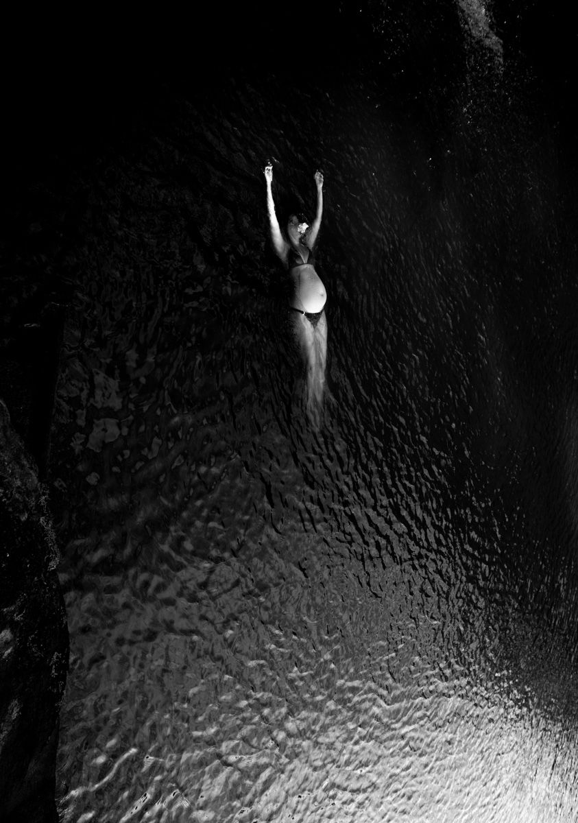 Black and white aerial view of a woman in bikini swimming with arms raised in dark rippling water.