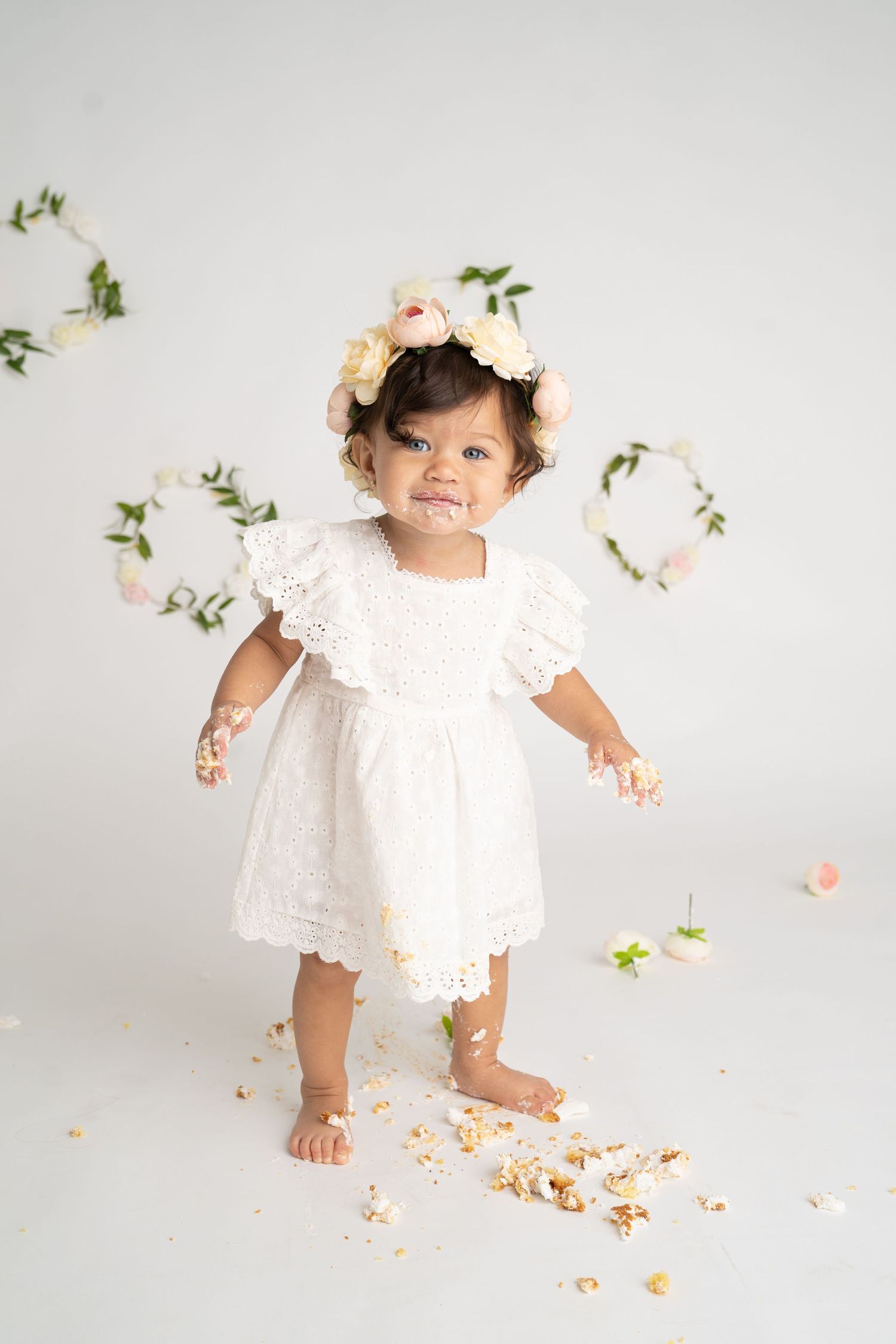 A happy toddler girl in a white lace dress and floral crown stands amid cake smash remnants on white backdrop.