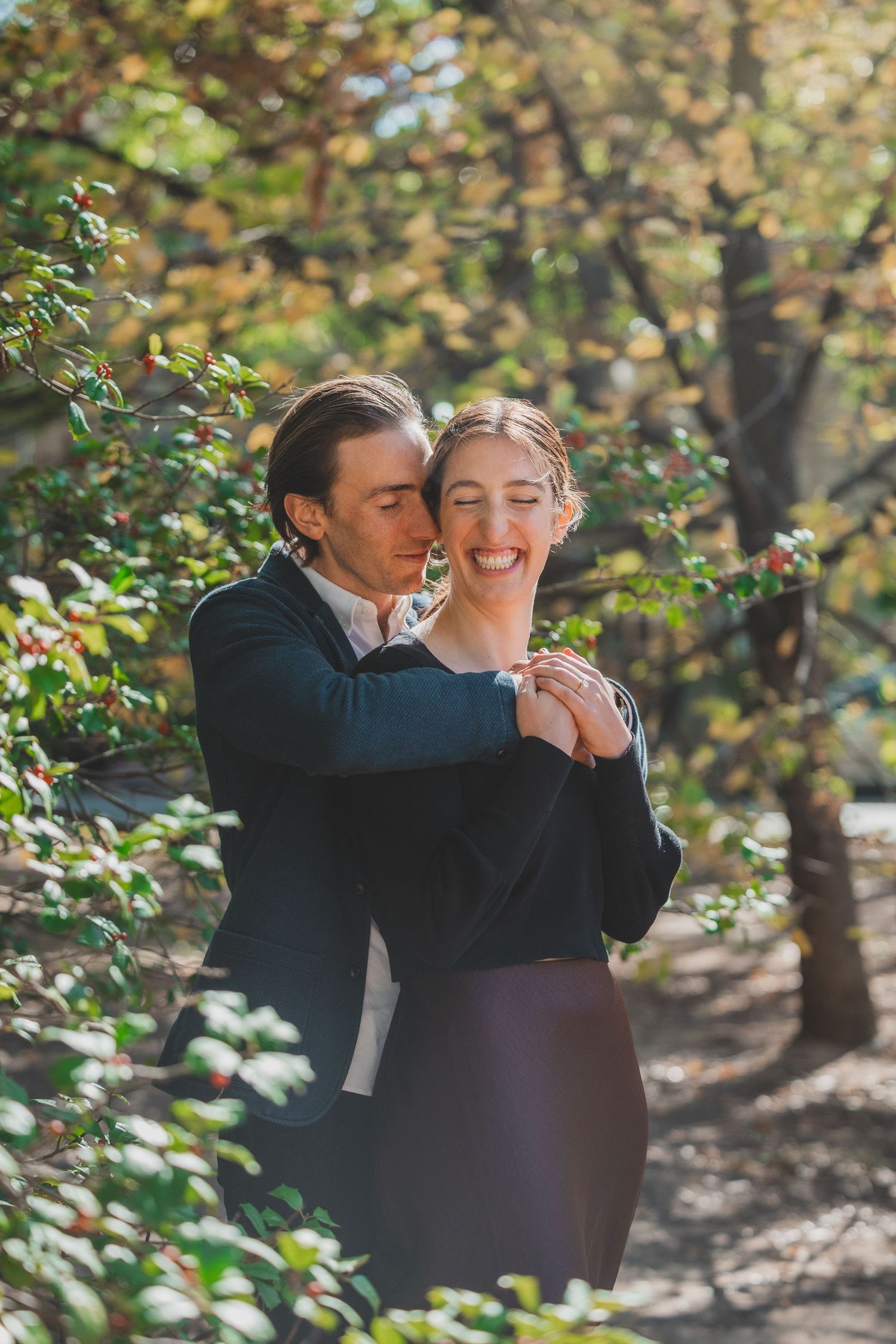 A couple embraces outdoors among autumn trees, the man hugging the woman from behind as she laughs joyfully.
