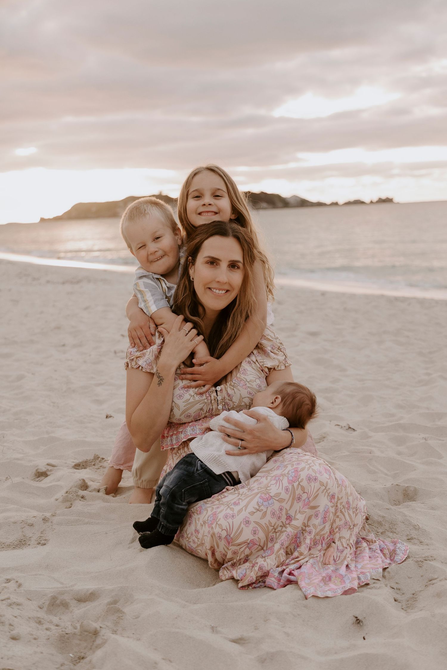 Family of four sitting on rocky beach at golden sunset with lush green hillside backdrop.