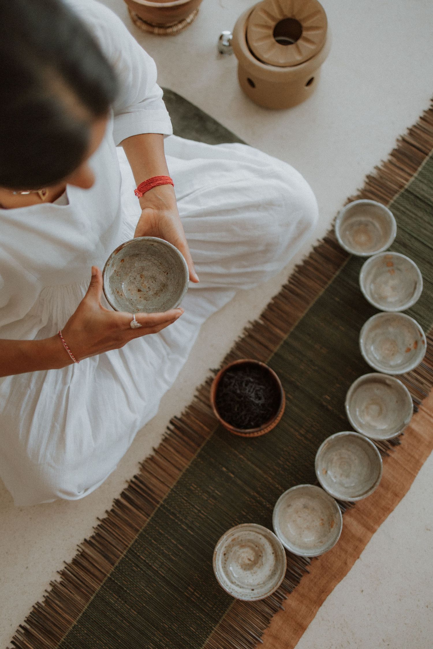 Woman in white seated cross-legged holding ceramic bowl during traditional tea ceremony with rustic cups arranged on mat.