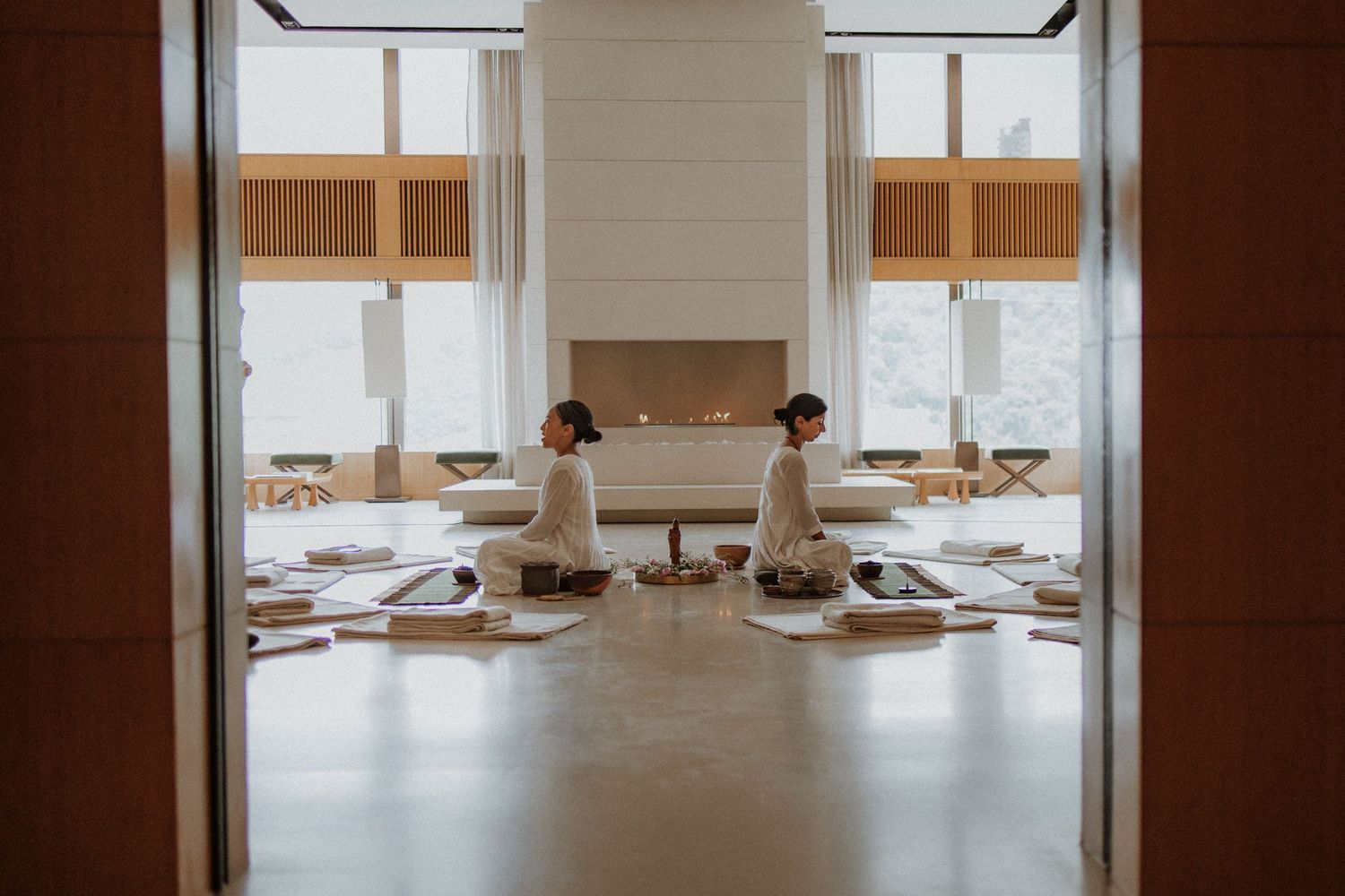 Two people in white robes sit facing each other in a serene spa meditation room with large windows and natural light.