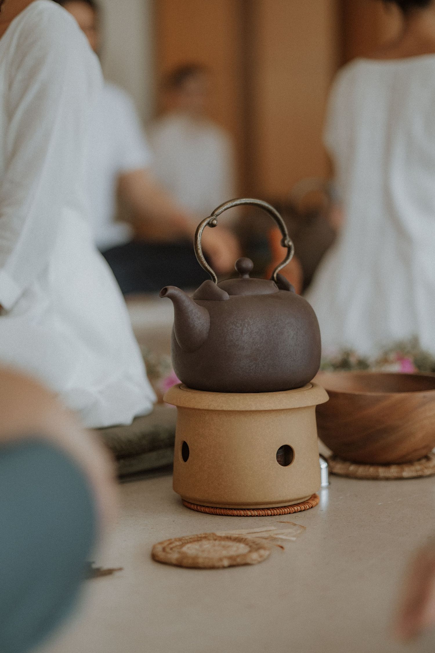 Dark clay teapot resting on a ceramic warmer during a traditional tea ceremony with people in white clothing.