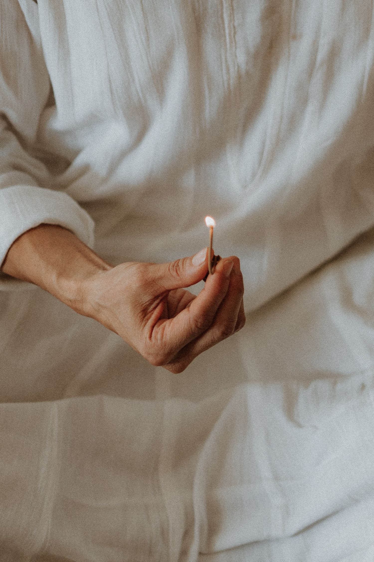 A hand in white linen clothing holds a lit match with a small flame against a soft white fabric background.