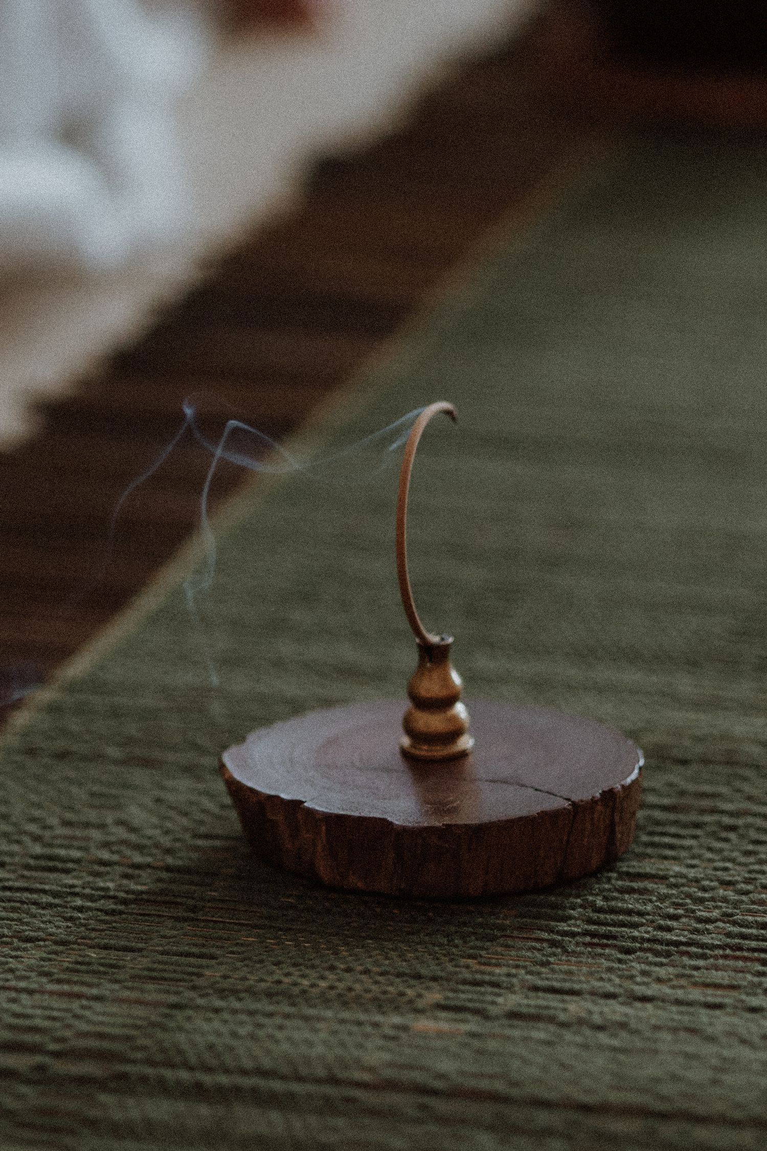A brass incense holder on a wooden slice sits on a green textile, with delicate smoke curling upward.