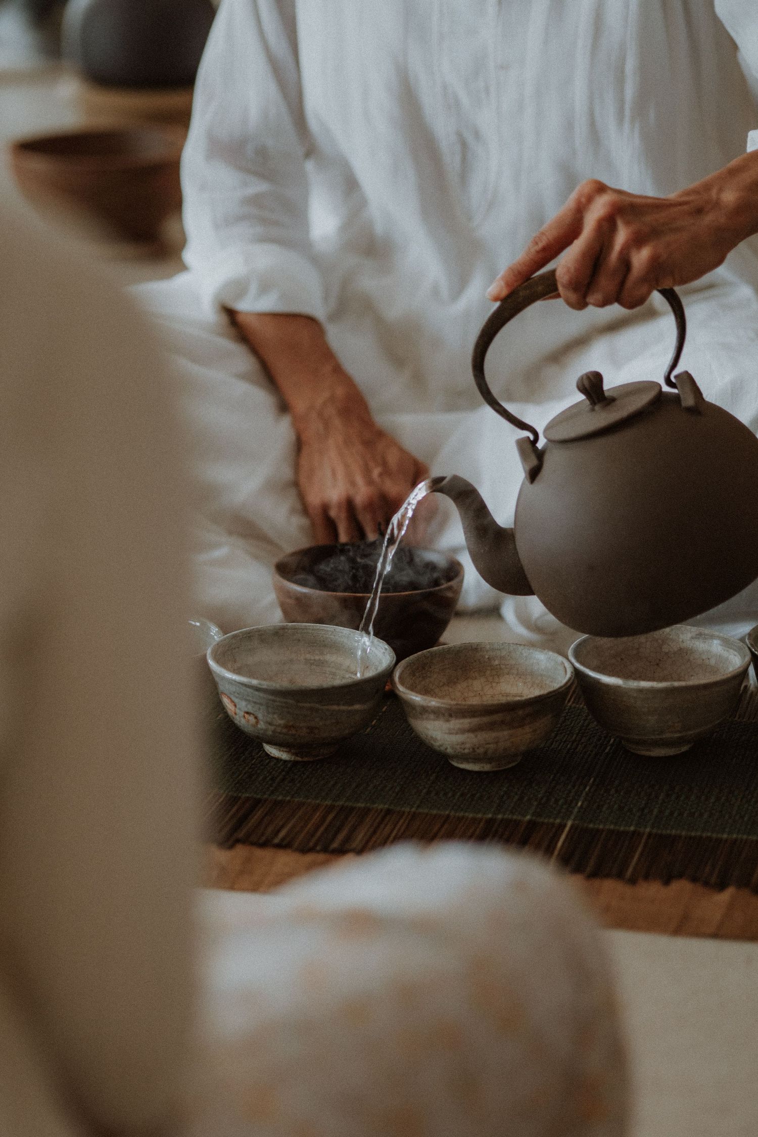 Person in white linen pouring hot water from dark clay teapot into ceramic bowls during traditional tea ceremony.