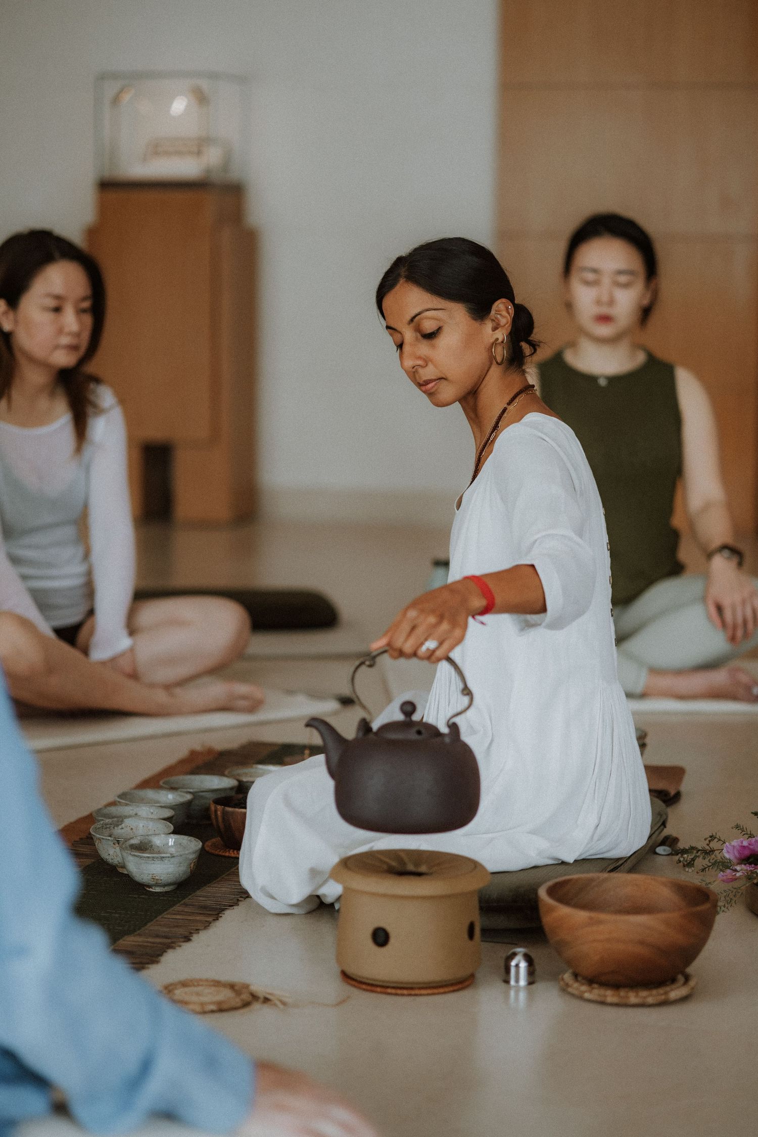 Woman in white performs traditional tea ceremony while guests meditate cross-legged in a serene studio setting.