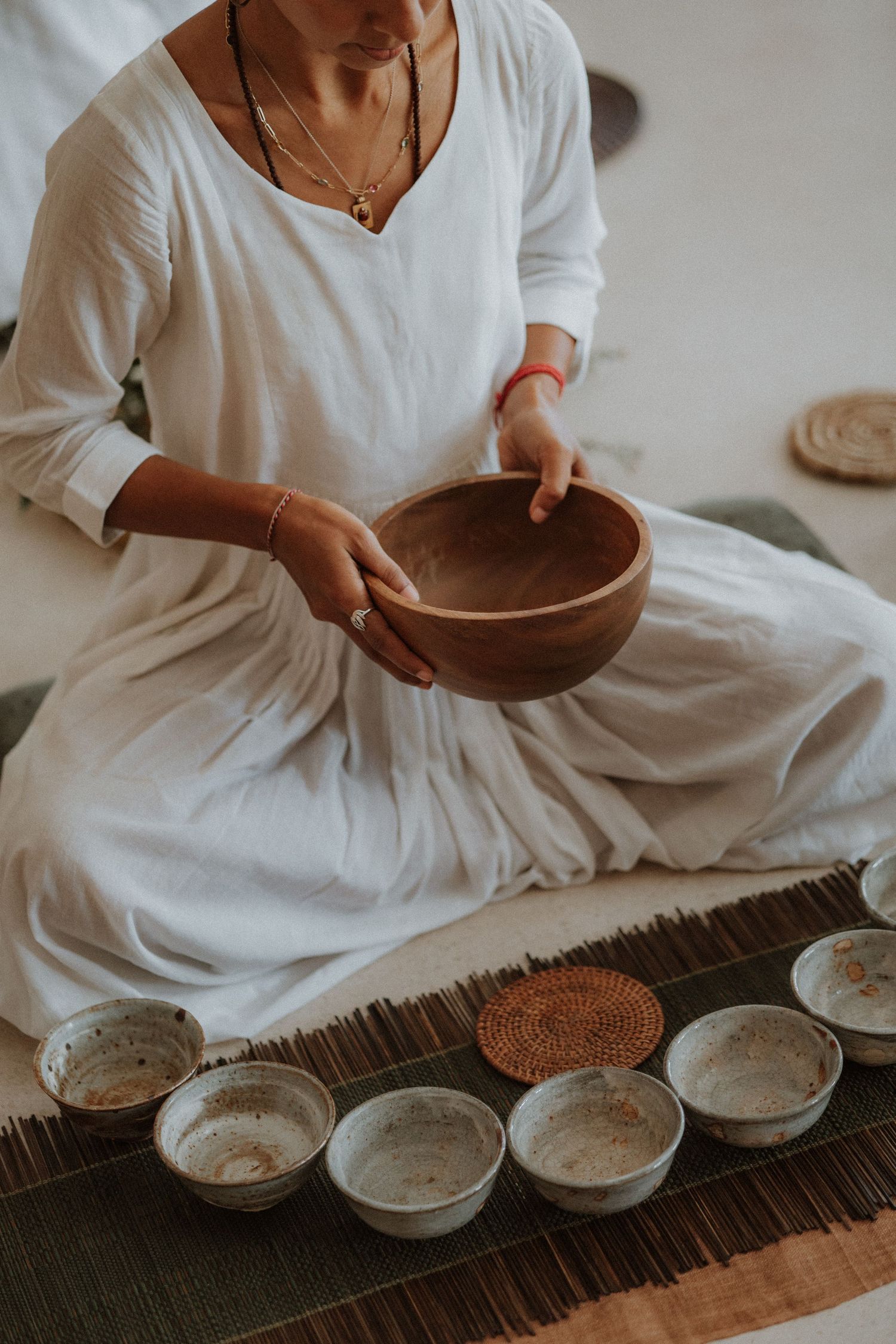 Woman in white dress sitting cross-legged holding wooden bowl with ceramic bowls arranged on mat during meditation ceremony.