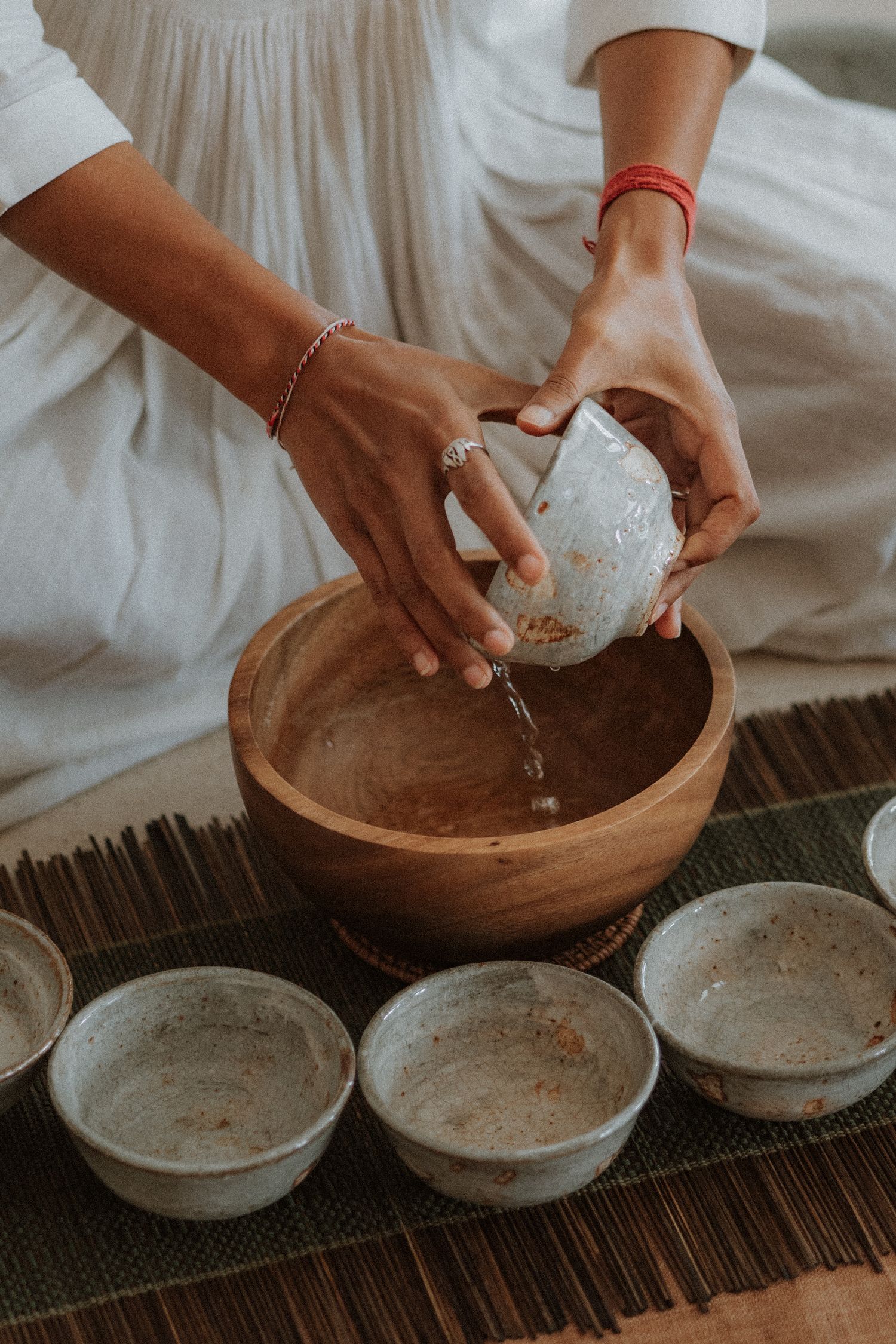 Woman in white dress pouring water from ceramic bowl into wooden bowl during traditional tea ceremony ritual.