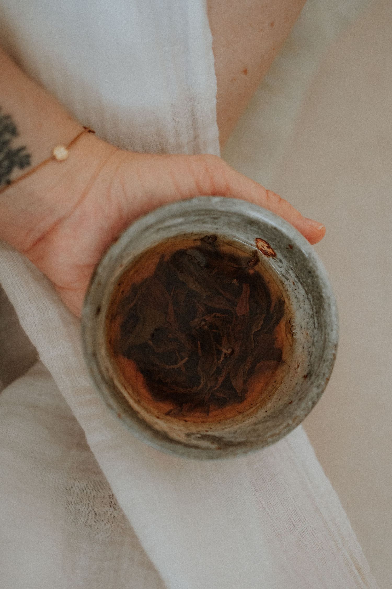 Hand holding a rustic ceramic bowl with dark tea leaves, wearing a gold bracelet, draped in white linen fabric.