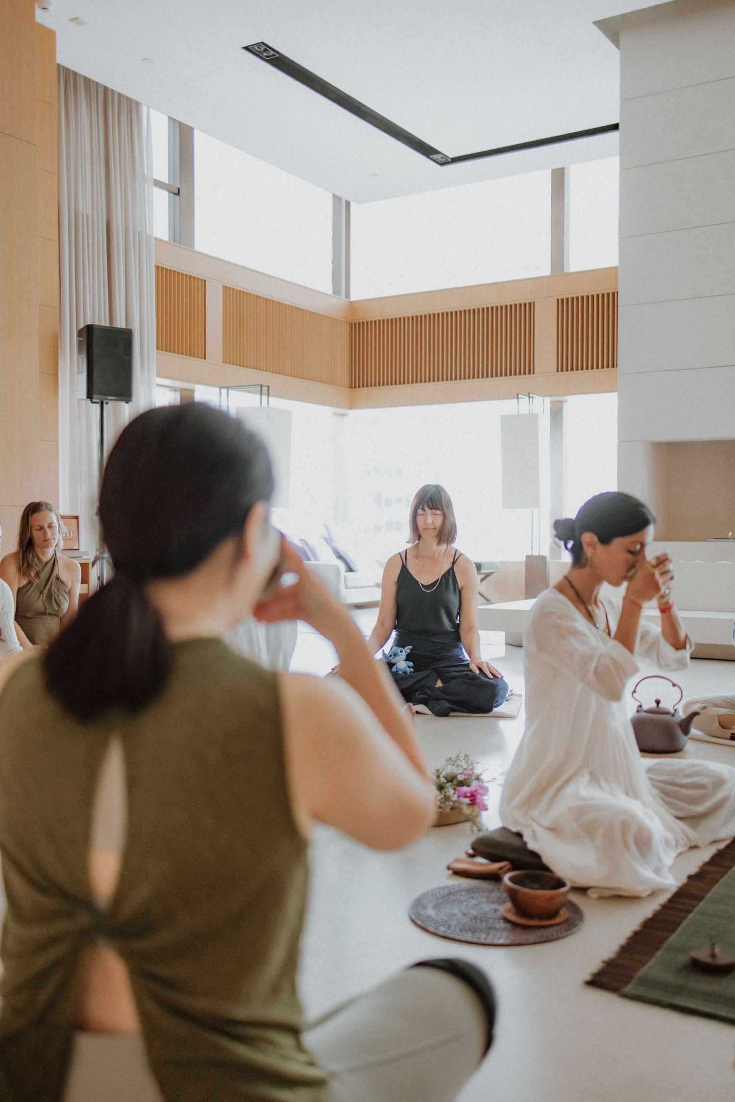 Women participate in a serene indoor tea ceremony and meditation session in a modern minimalist space with large windows.