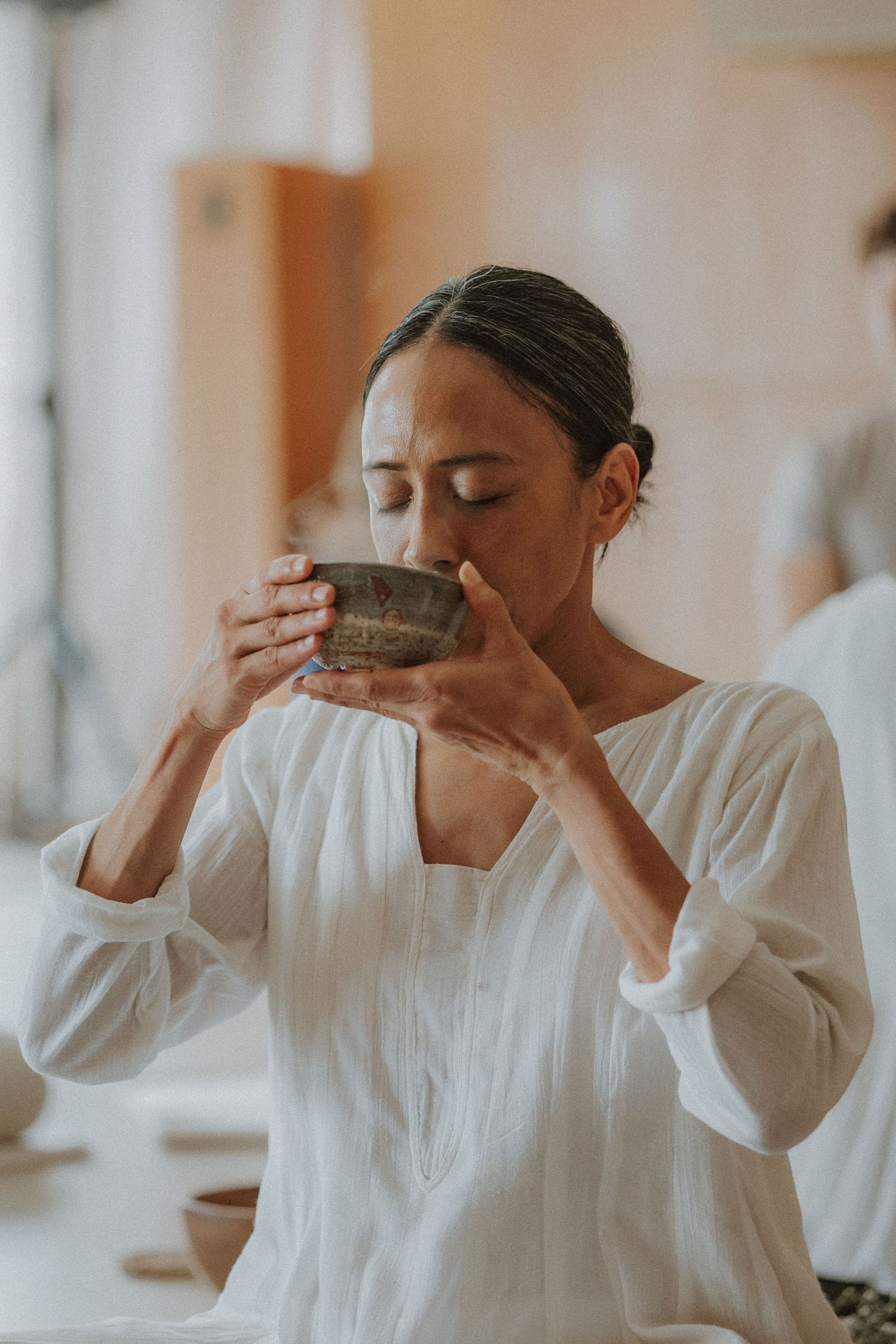 Woman in white linen top sipping from a rustic ceramic bowl with eyes closed in a serene studio setting.