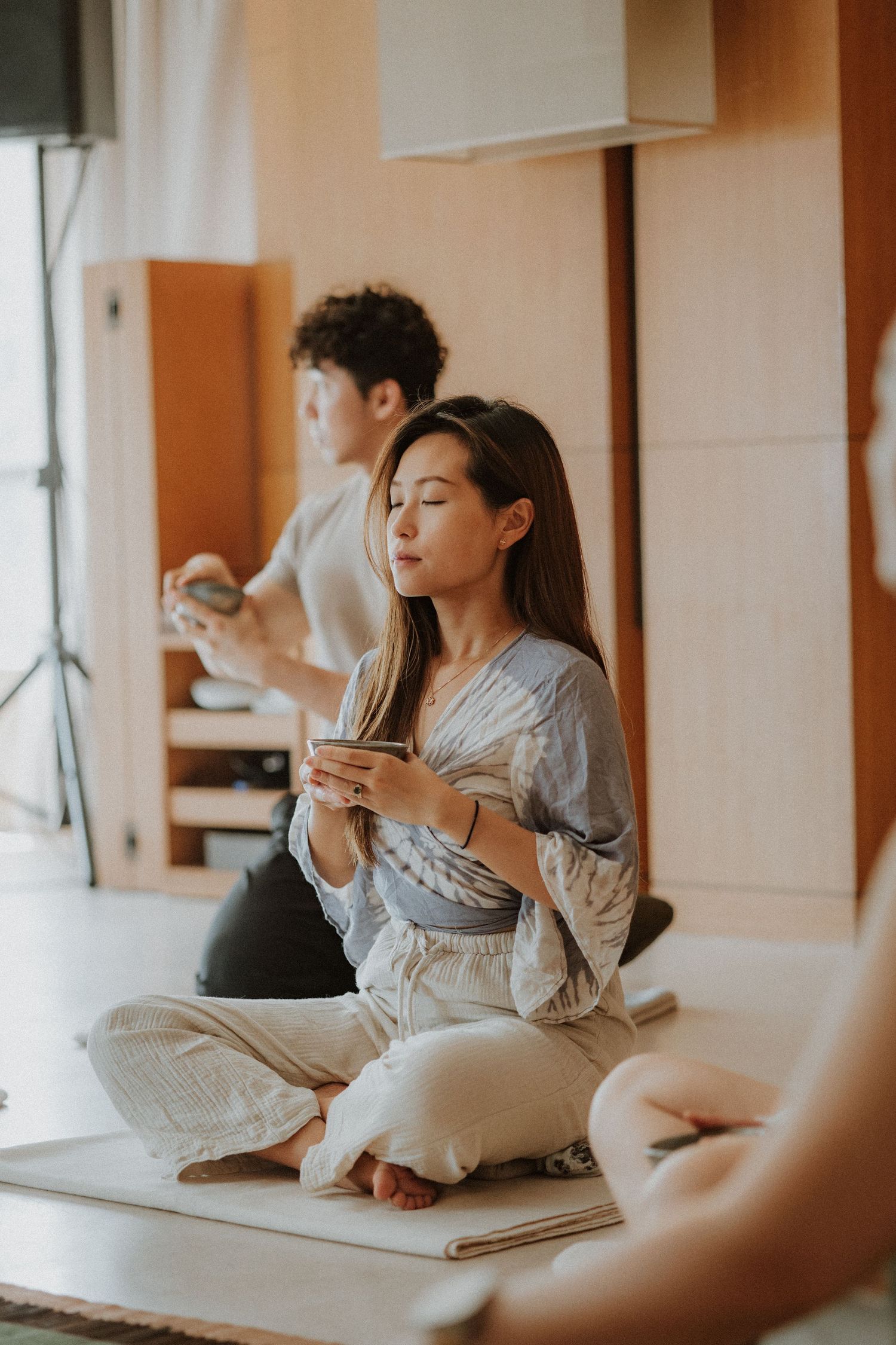 Woman sitting cross-legged with eyes closed, holding a tea bowl during a mindful Japanese tea ceremony indoors.