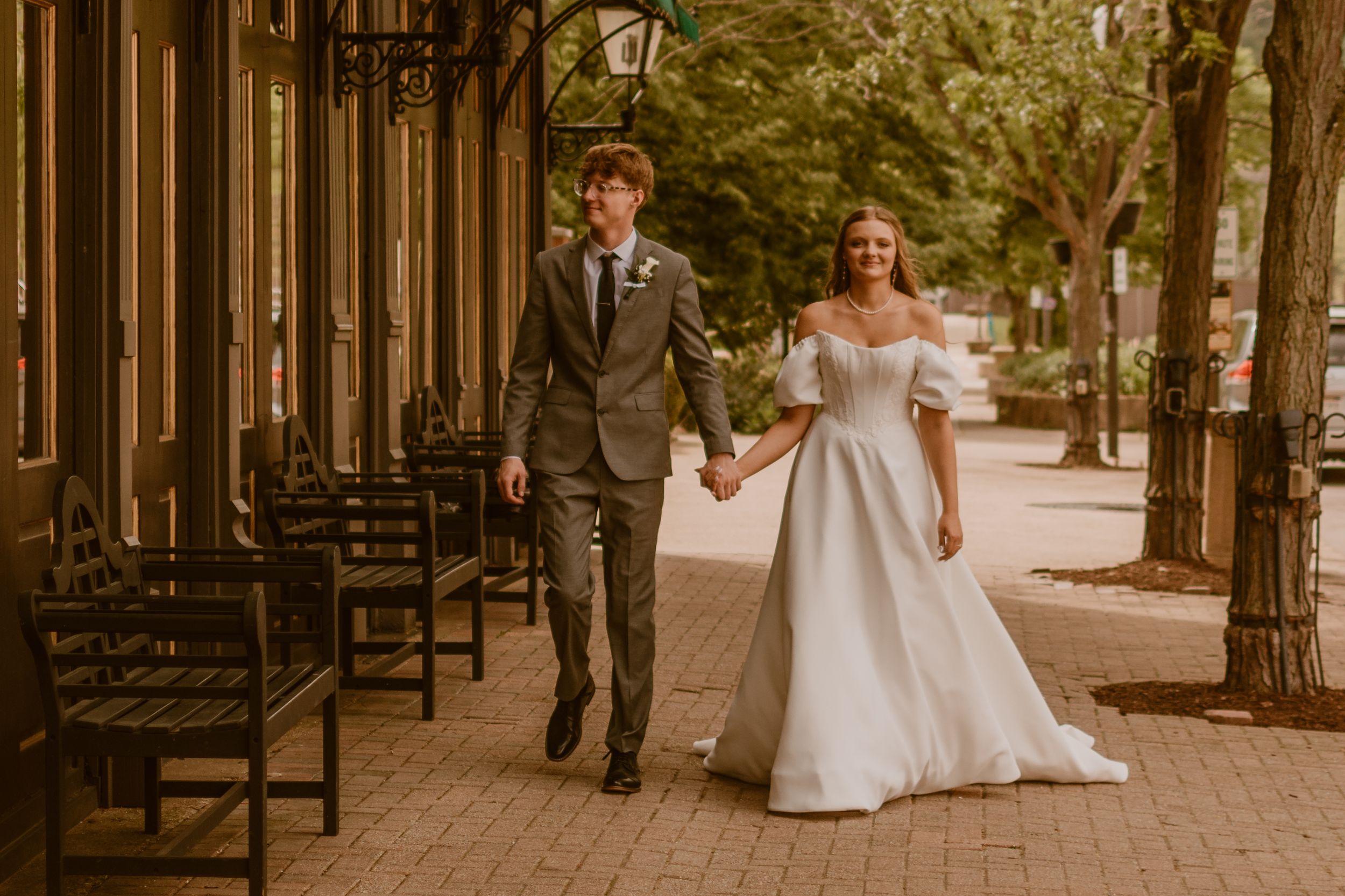 Bride and groom walk hand in hand on a brick sidewalk, she in a white gown, he in a gray suit.