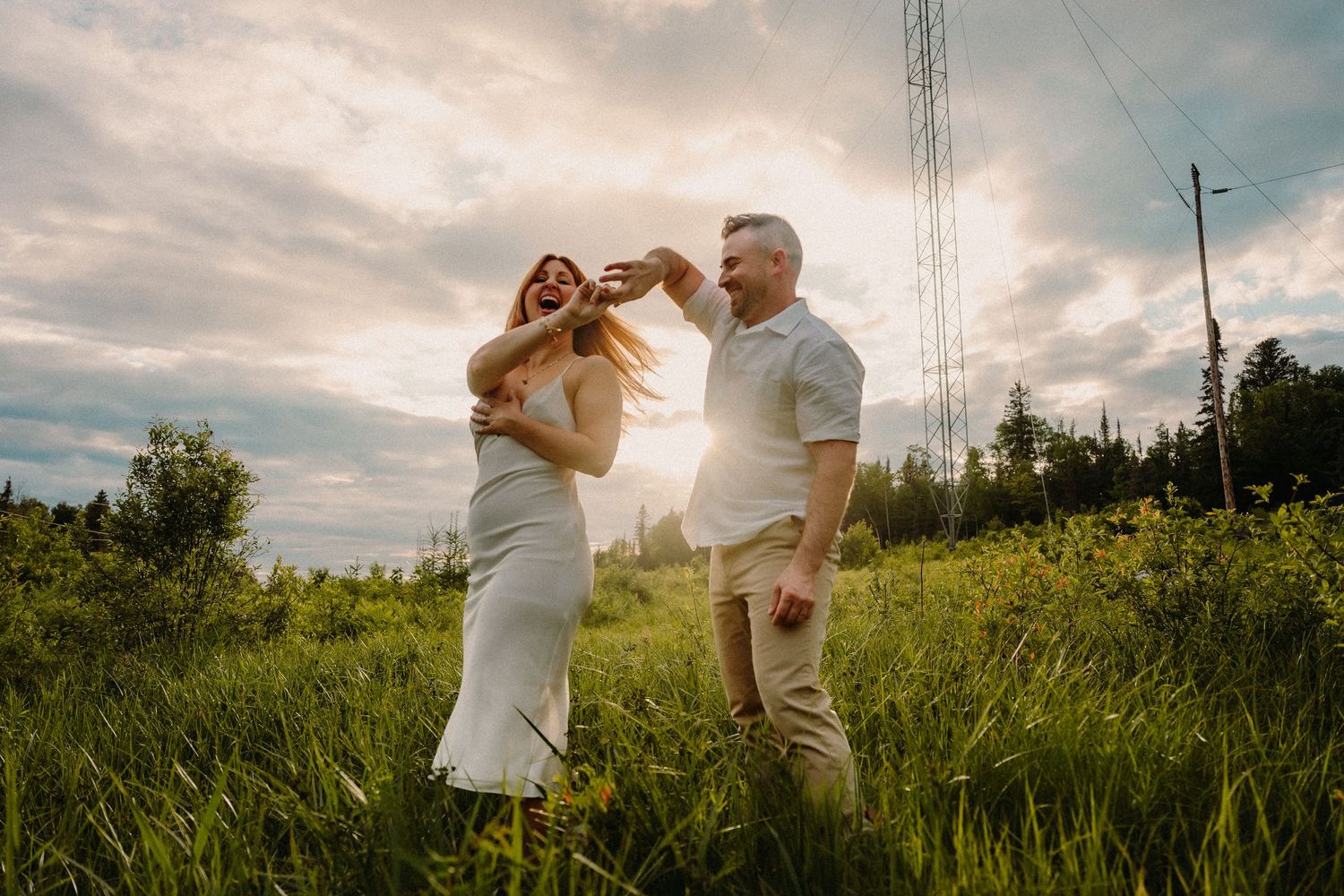 Couple dancing joyfully in a sunny green field at golden hour, woman in white dress, man in casual attire.