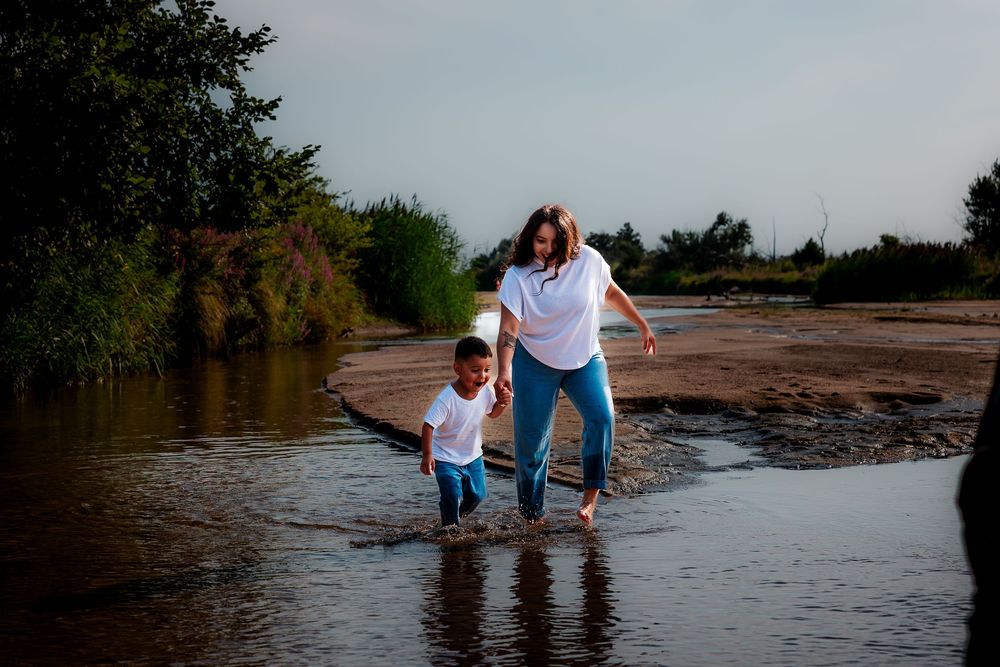 A mother and child splash through shallow water near a riverbank, both wearing white shirts and jeans on a cloudy day.