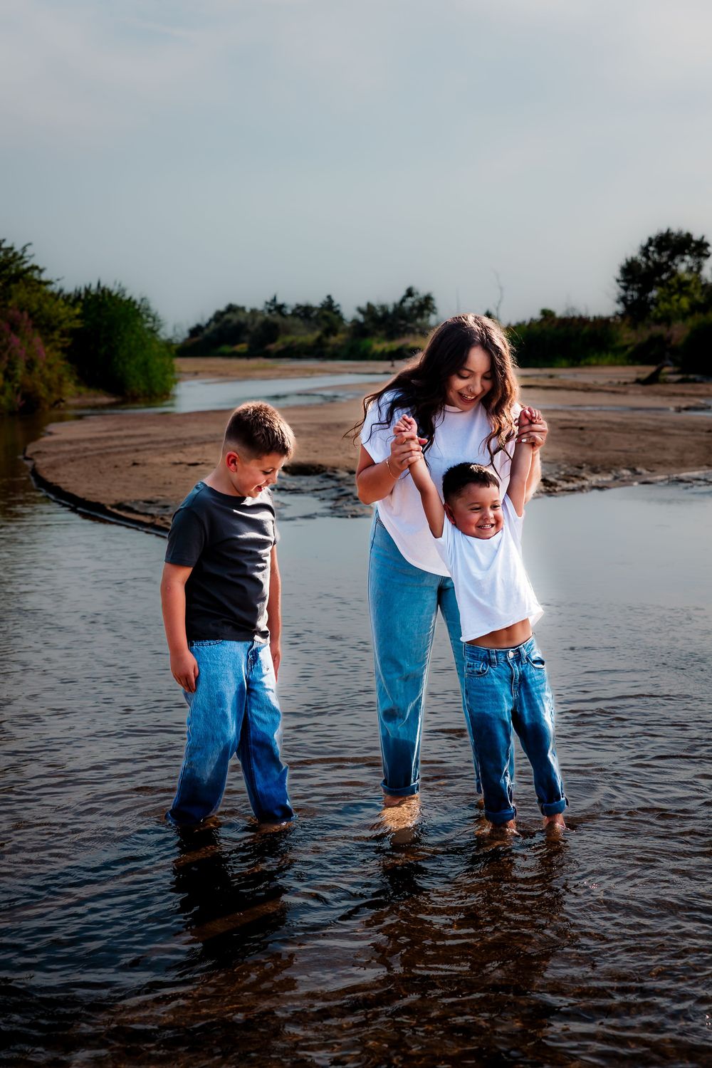 A mother and two young boys wade barefoot in shallow water at a sandy beach, all wearing jeans and casual tops.