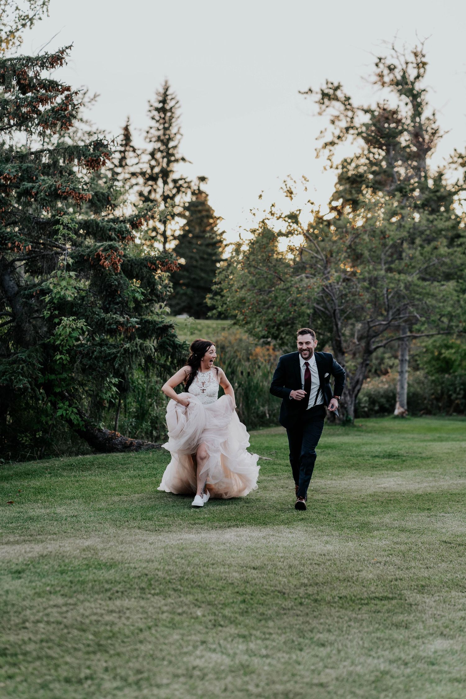 A joyful bride and groom run across a lush green lawn surrounded by tall evergreen trees at dusk.