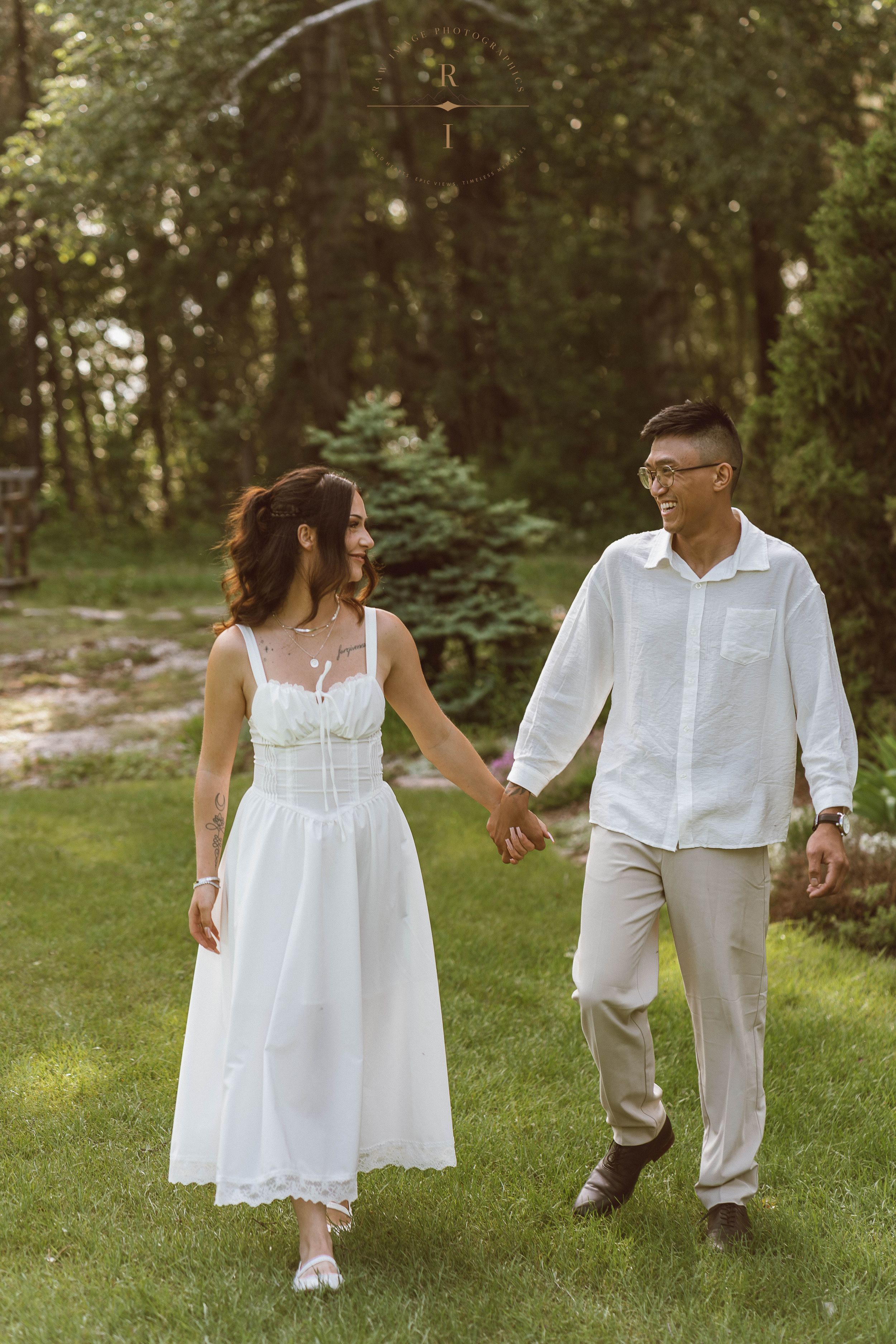 A couple in white attire holds hands while walking on green grass in a lush garden setting.