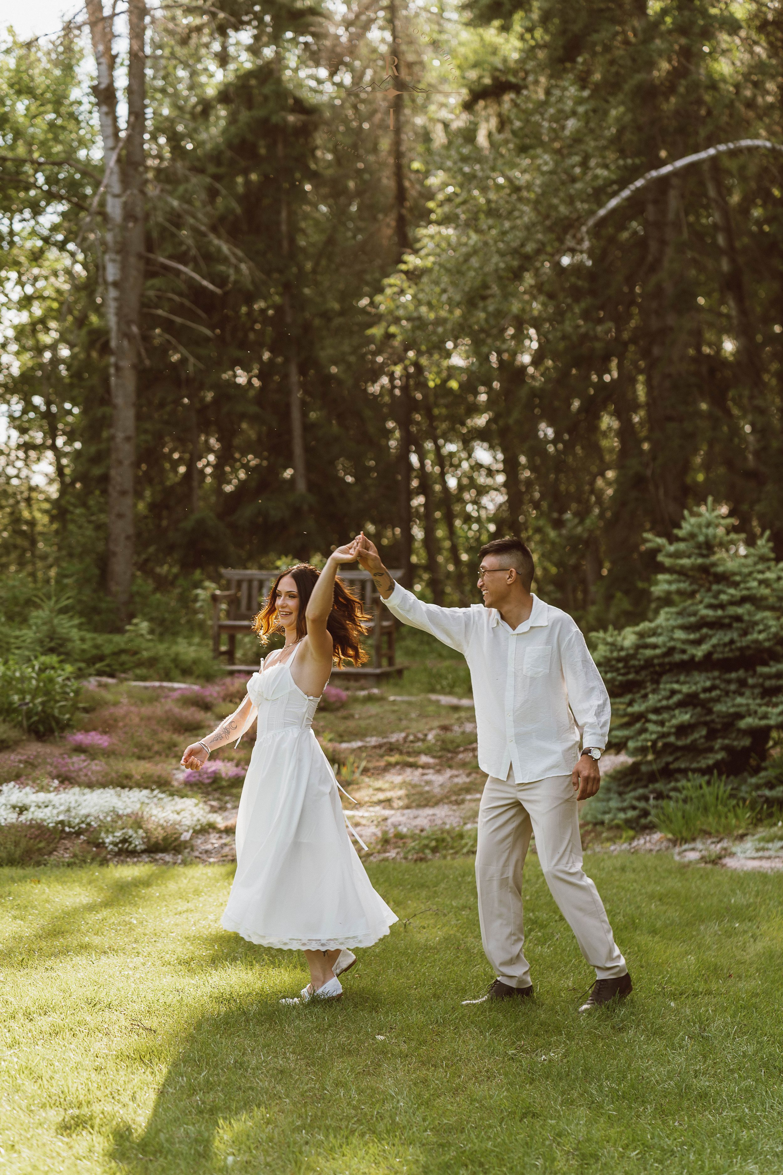 Couple dancing outdoors on green lawn in garden, woman in white dress being twirled by man in white shirt.