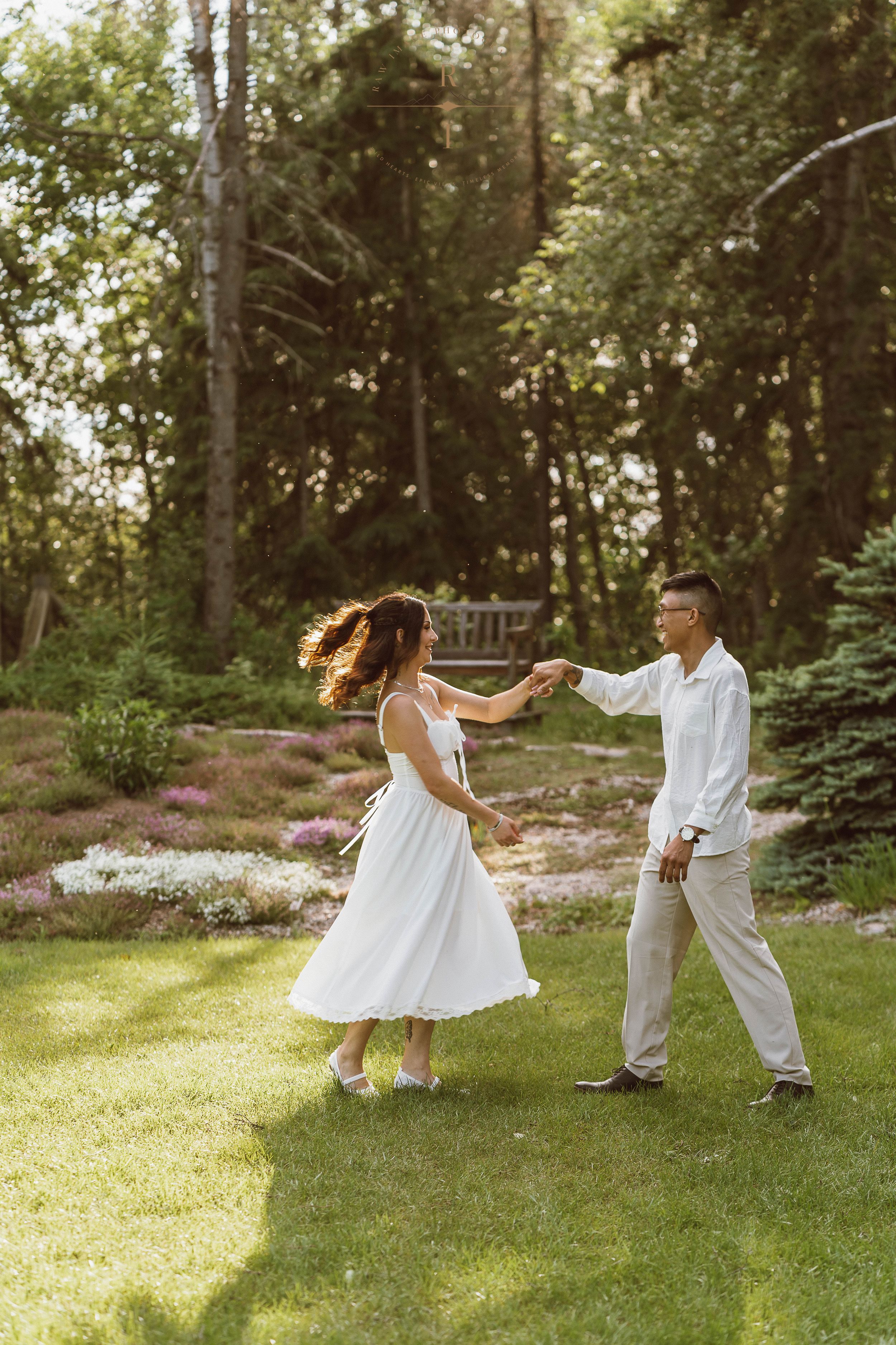 Couple dancing together on lush green lawn surrounded by blooming garden and tall trees in warm sunlight.