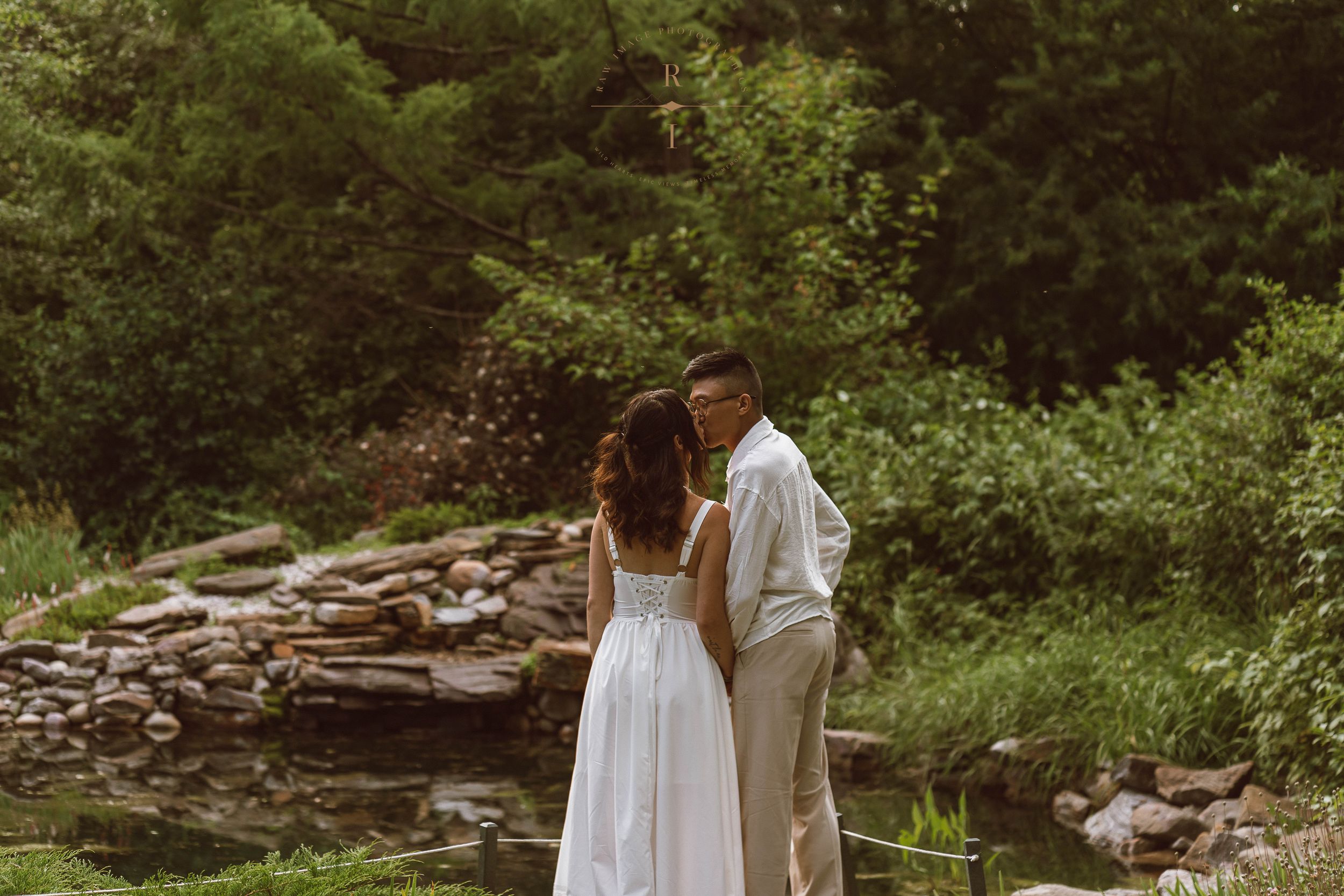 Bride and groom standing hand in hand near a serene stone-lined pond surrounded by lush green trees.