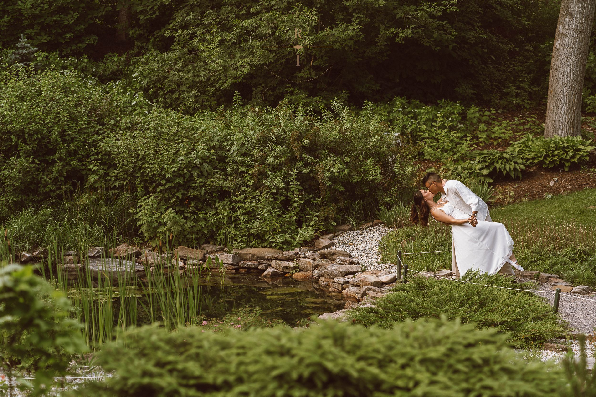 Bride in white dress kneeling by a serene garden pond surrounded by lush green plants and trees.