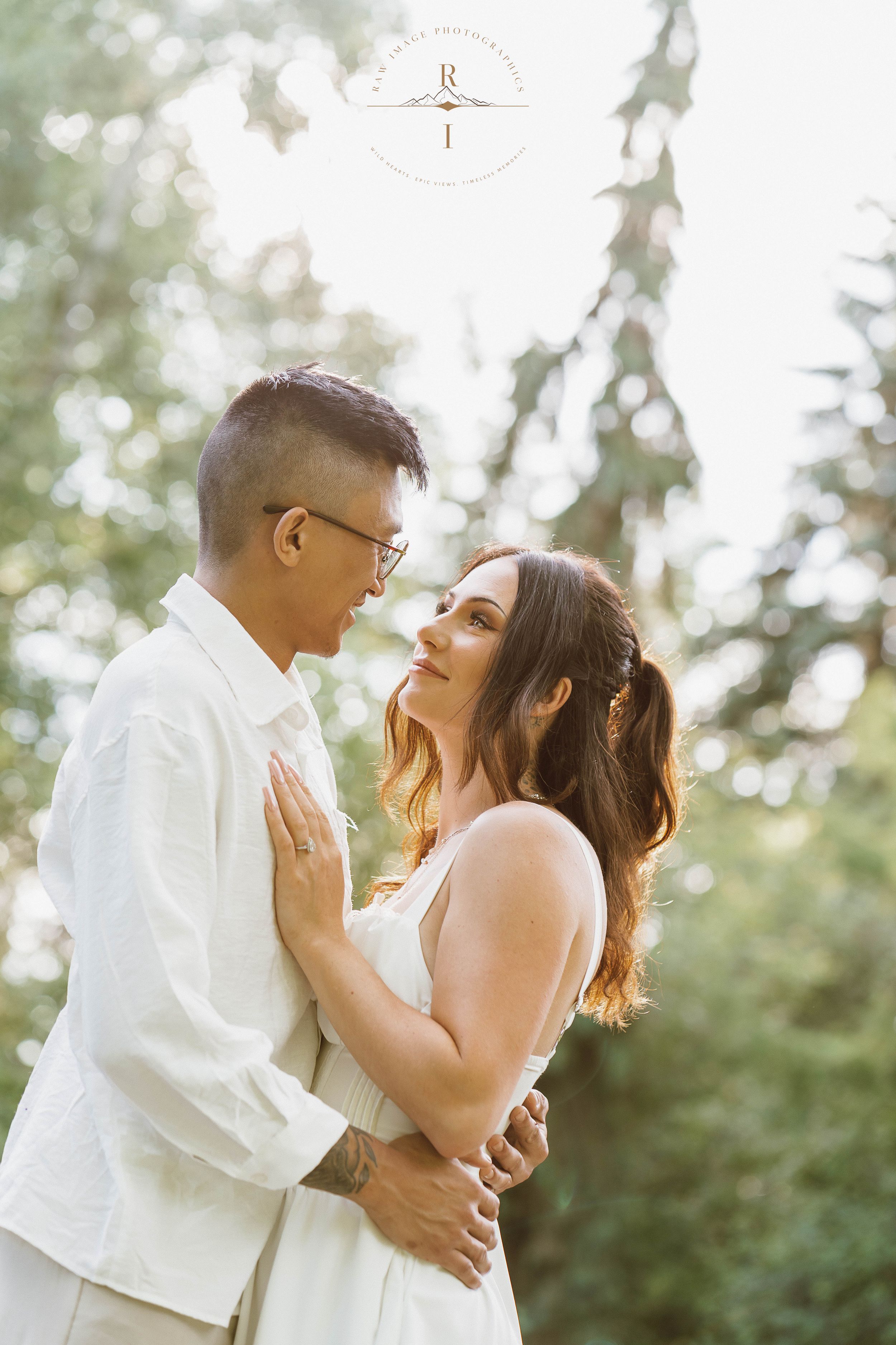 Couple in white outfits embracing outdoors, sharing a romantic moment surrounded by lush green trees in soft natural light.