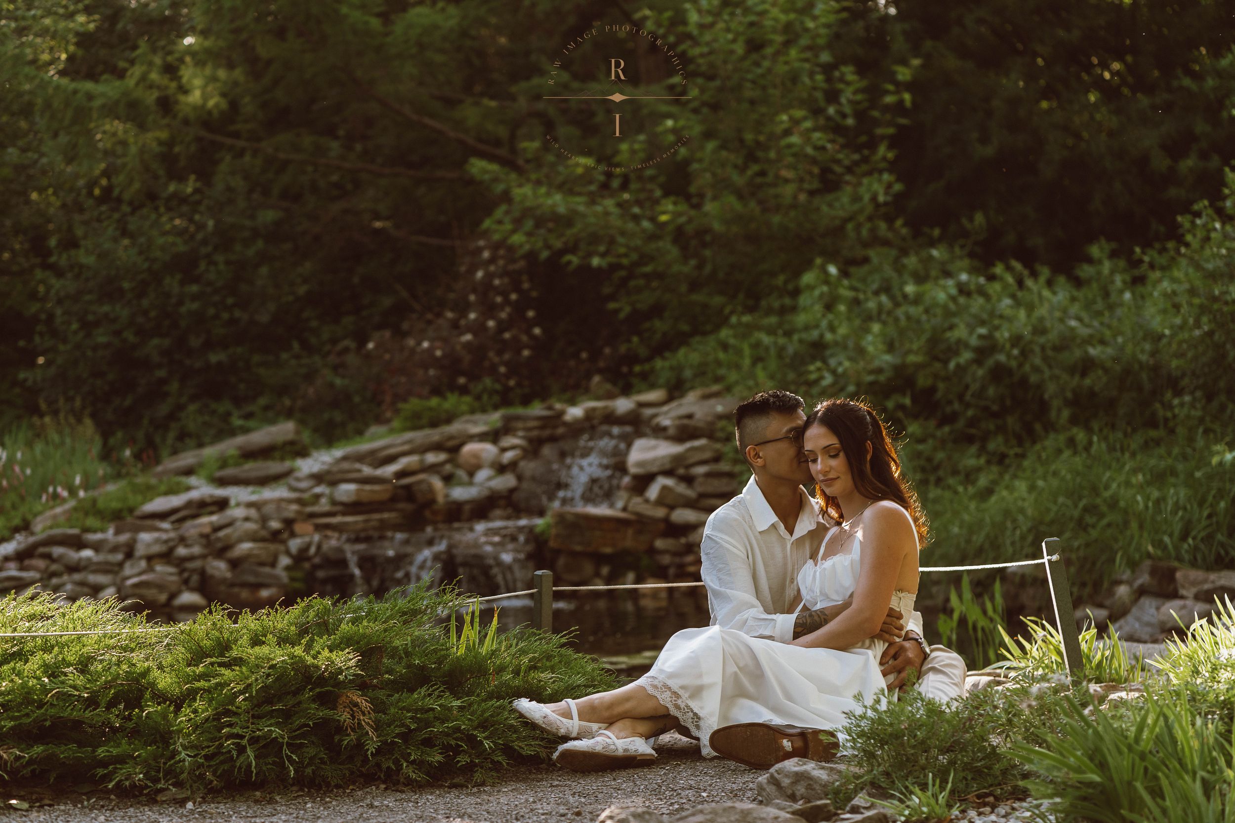 Couple in white outfits sitting together near a stone waterfall surrounded by lush green gardens.