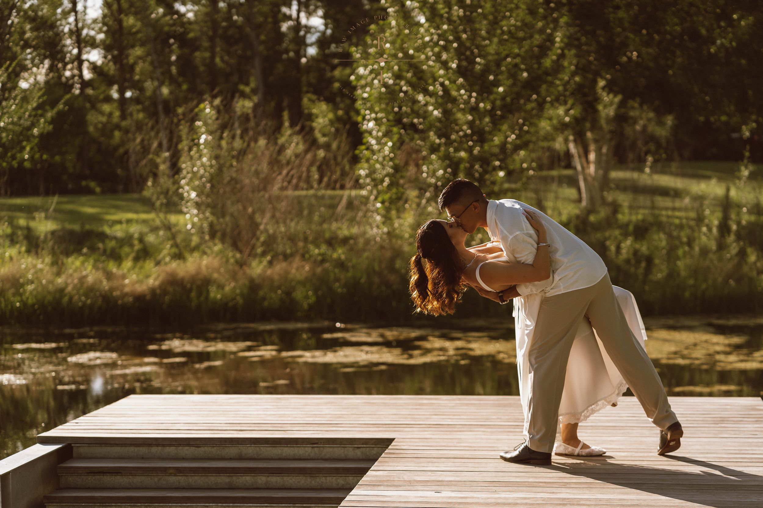 Couple sharing a romantic dip kiss on a wooden dock surrounded by lush greenery and a serene pond.
