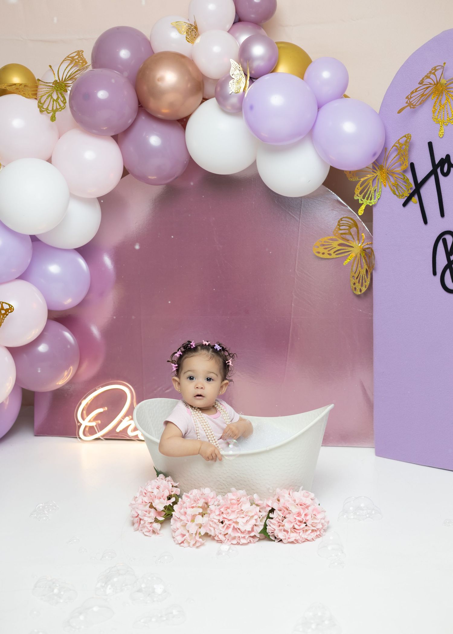 Baby girl in white tub surrounded by pink flowers and purple balloons at a first birthday celebration.