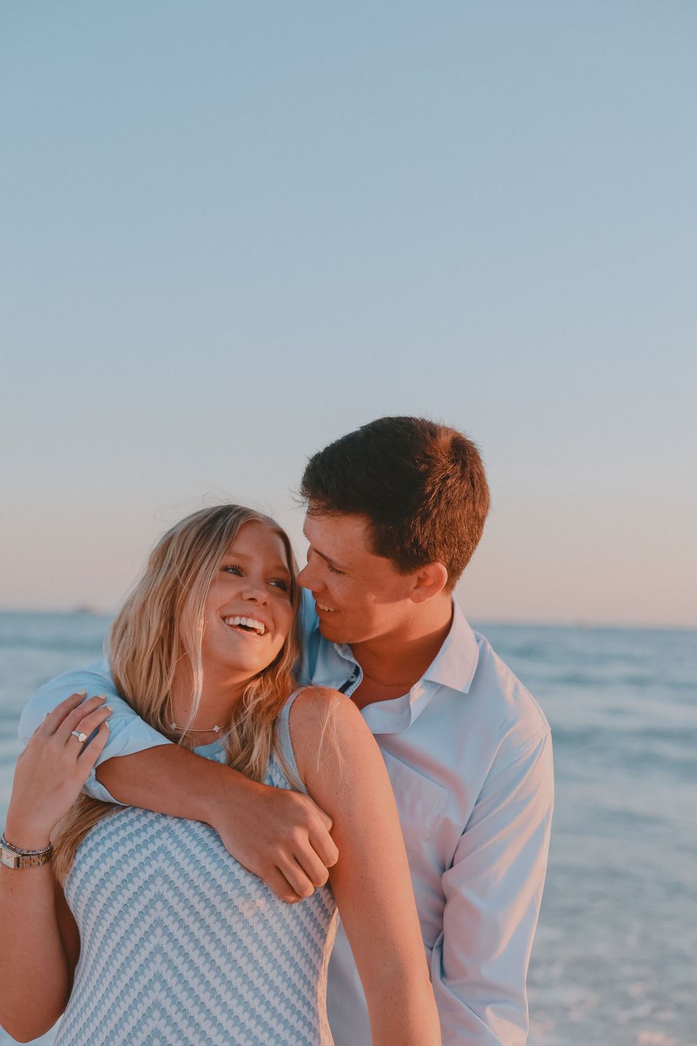 Happy engaged couple embracing on sandy beach during golden sunset with colorful cloudy sky.