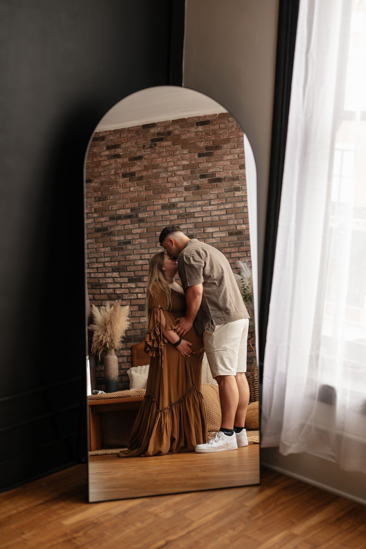 Couple kissing in reflection of arched floor mirror, brick wall background, woman in olive maxi dress, man in casual outfit.