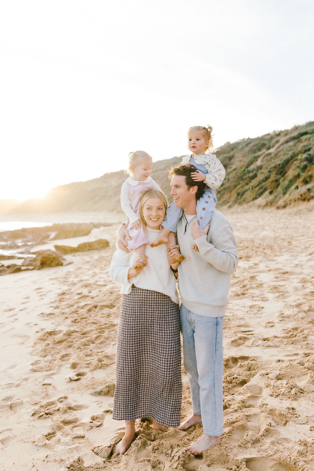 Family of four posing on a sandy beach at sunset, parents holding two young children, with rocky cliffs in the background.