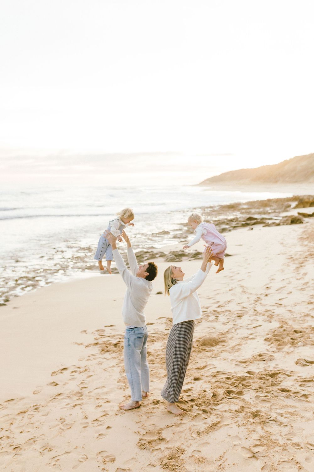 A joyful family lifting two toddlers in the air on a sunny beach with golden sand and ocean waves.