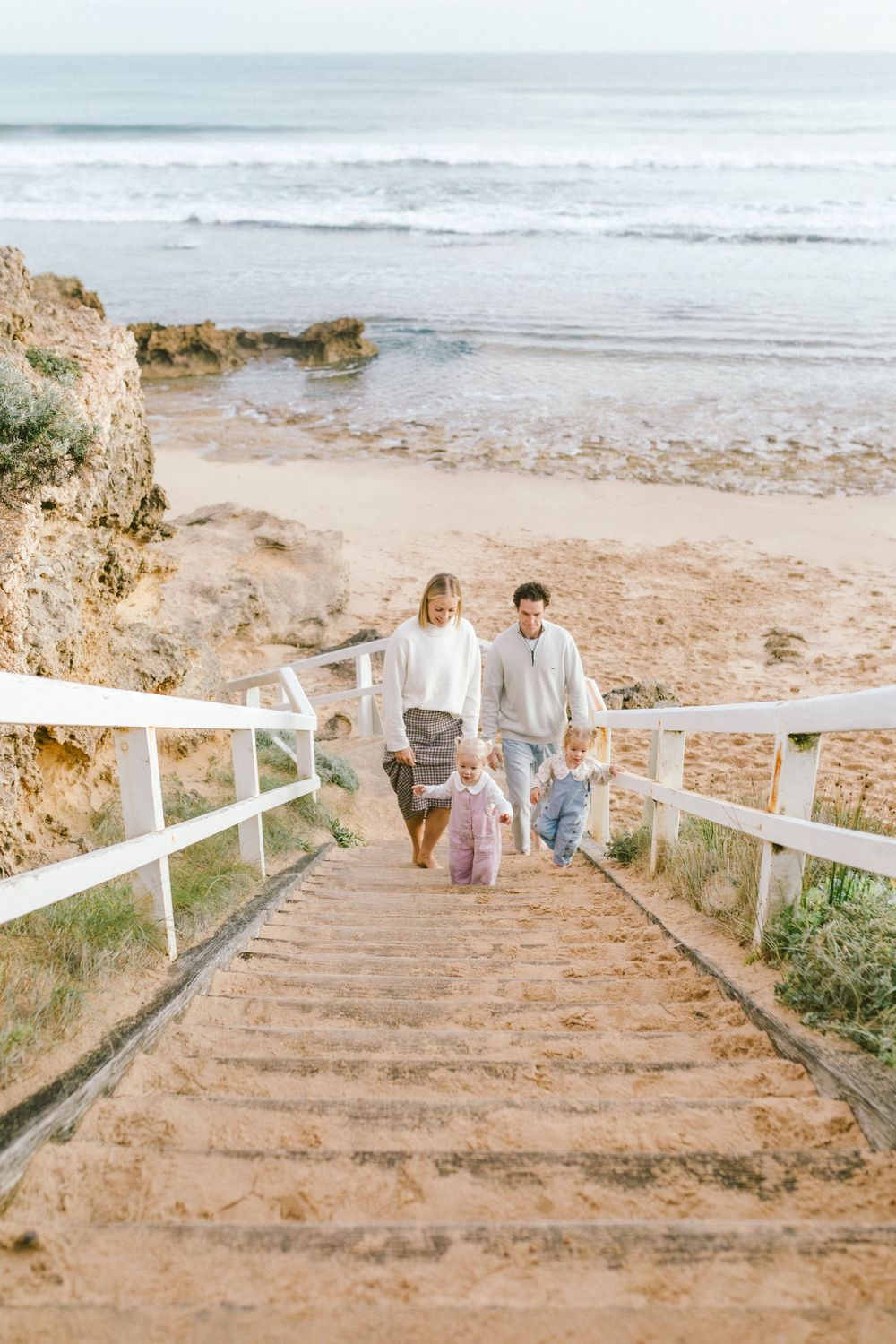 Family with two toddlers climbing sandy beach stairs with ocean waves and rocky cliffs in the background.