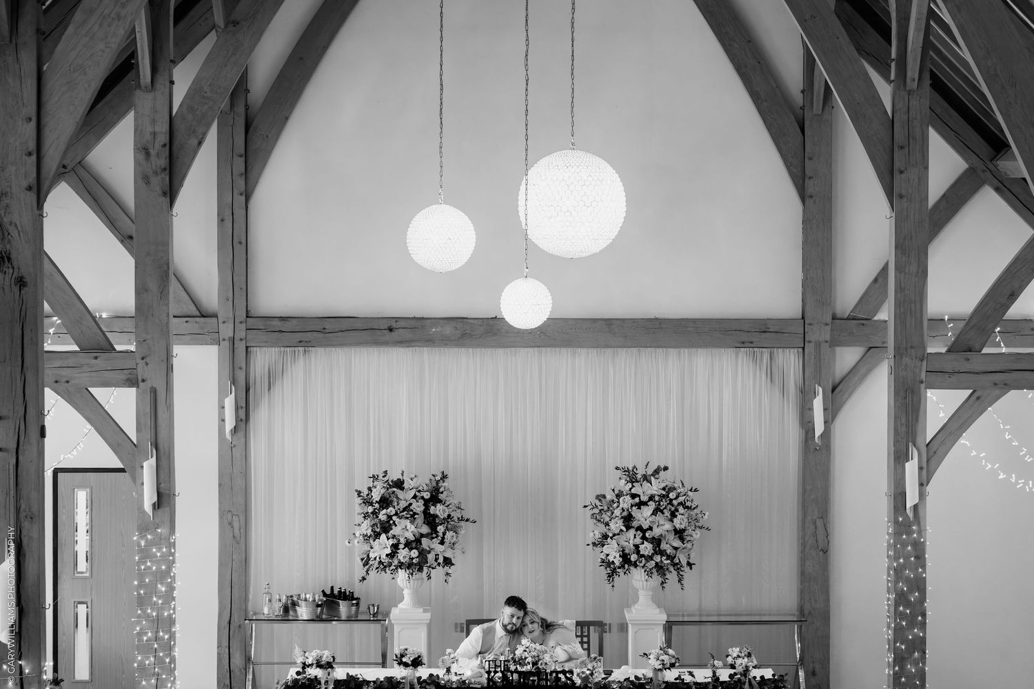 Elegant barn wedding reception with timber frame arches, globe pendant lights, and floral arrangements on the head table.