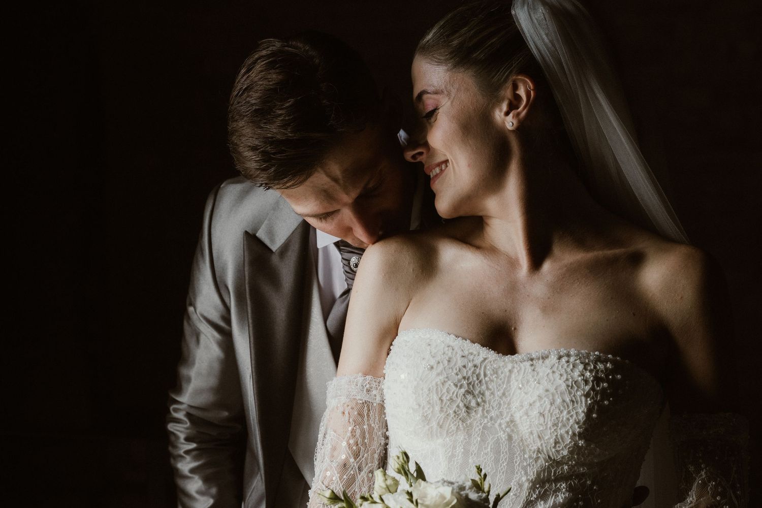 Bride and groom share an intimate moment, she smiles in a strapless lace gown with veil, holding a white bouquet.