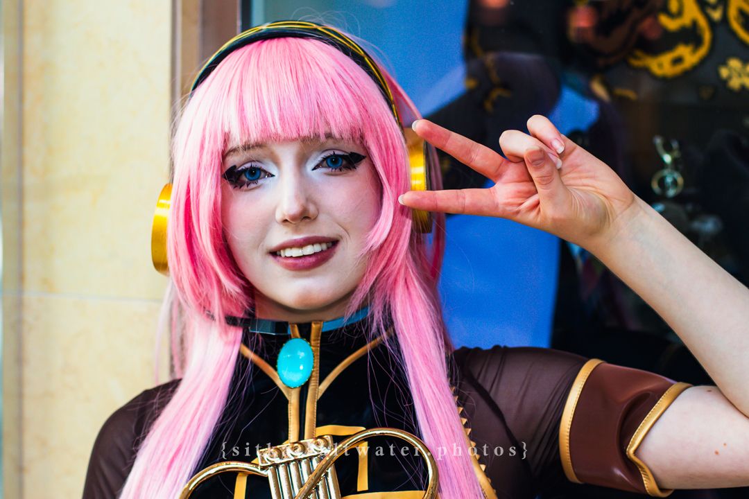 Person cosplaying with pink wig and headphones, making peace sign, wearing detailed costume at convention.