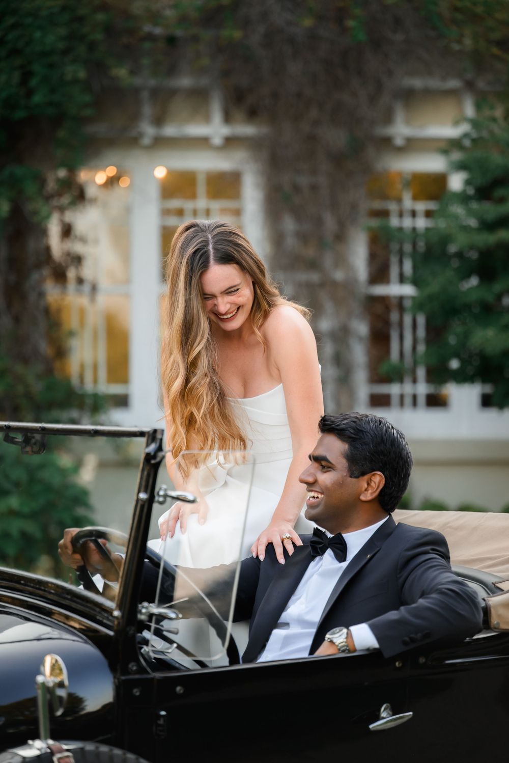 Bride in white strapless gown laughing with groom in black tuxedo seated in vintage convertible car outside elegant venue.