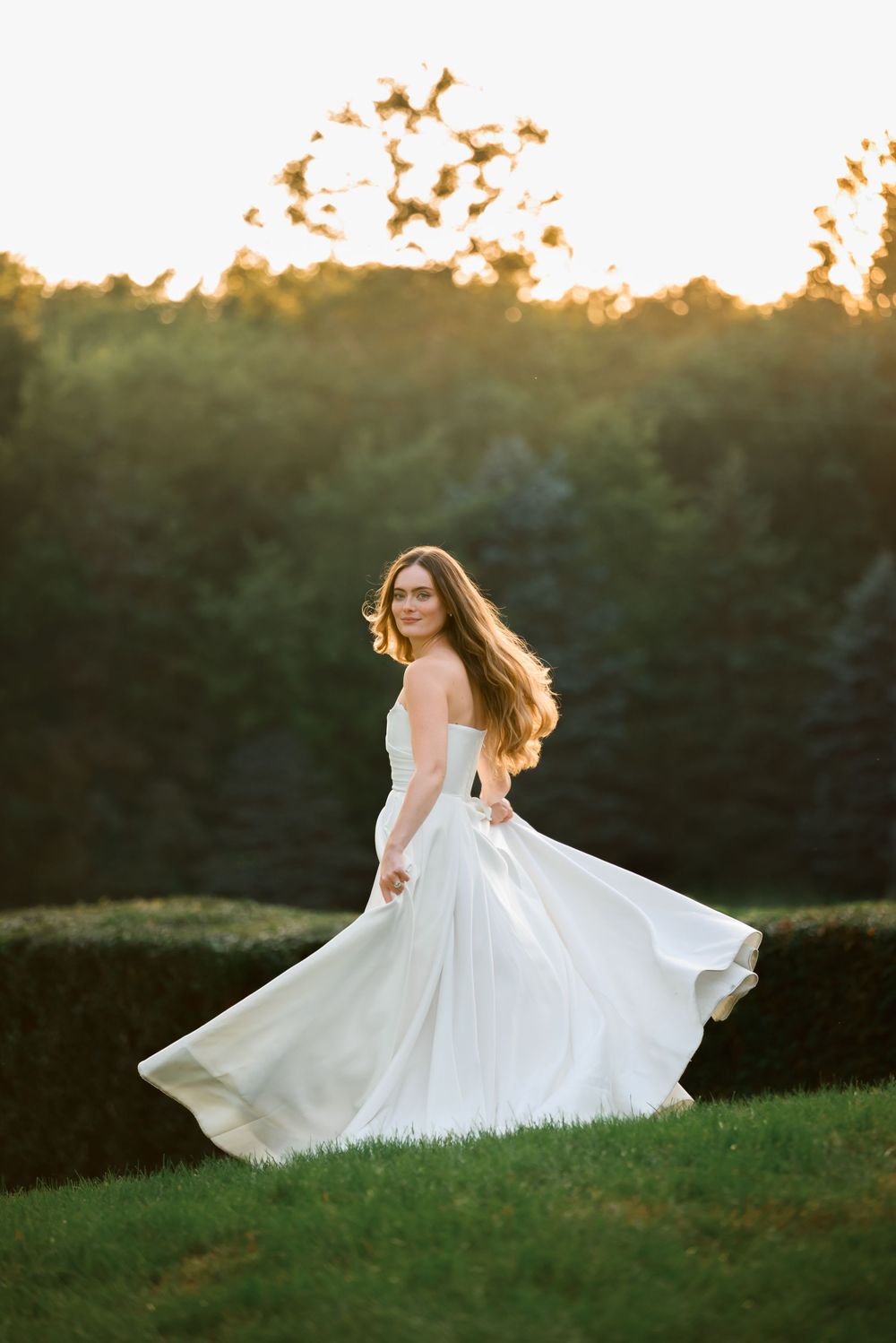 Bride in flowing white gown twirling outdoors at golden hour with lush green hedges and trees in background.