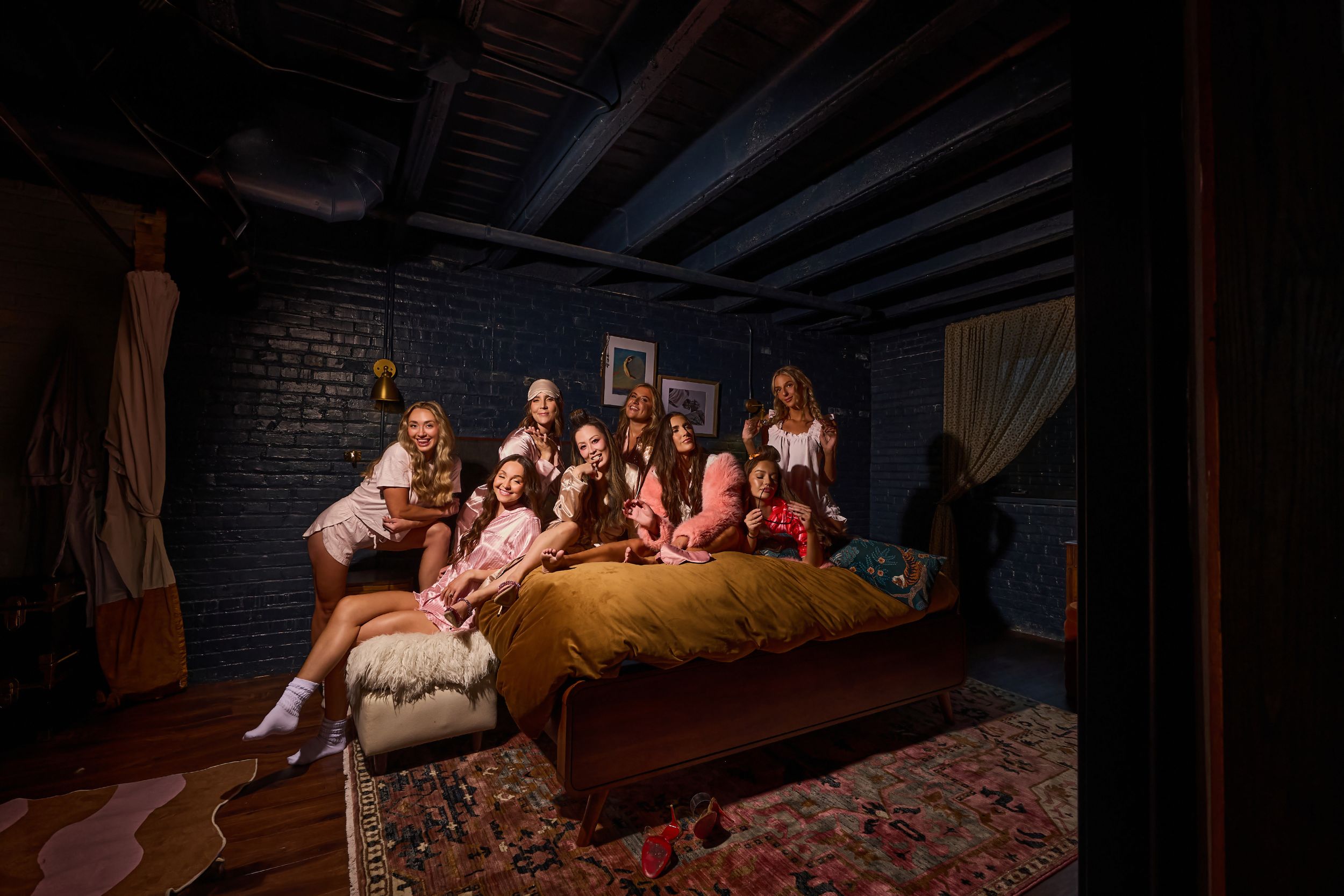 Group of women posing together on a bed in a cozy, dimly lit room with brick walls and wooden beams.