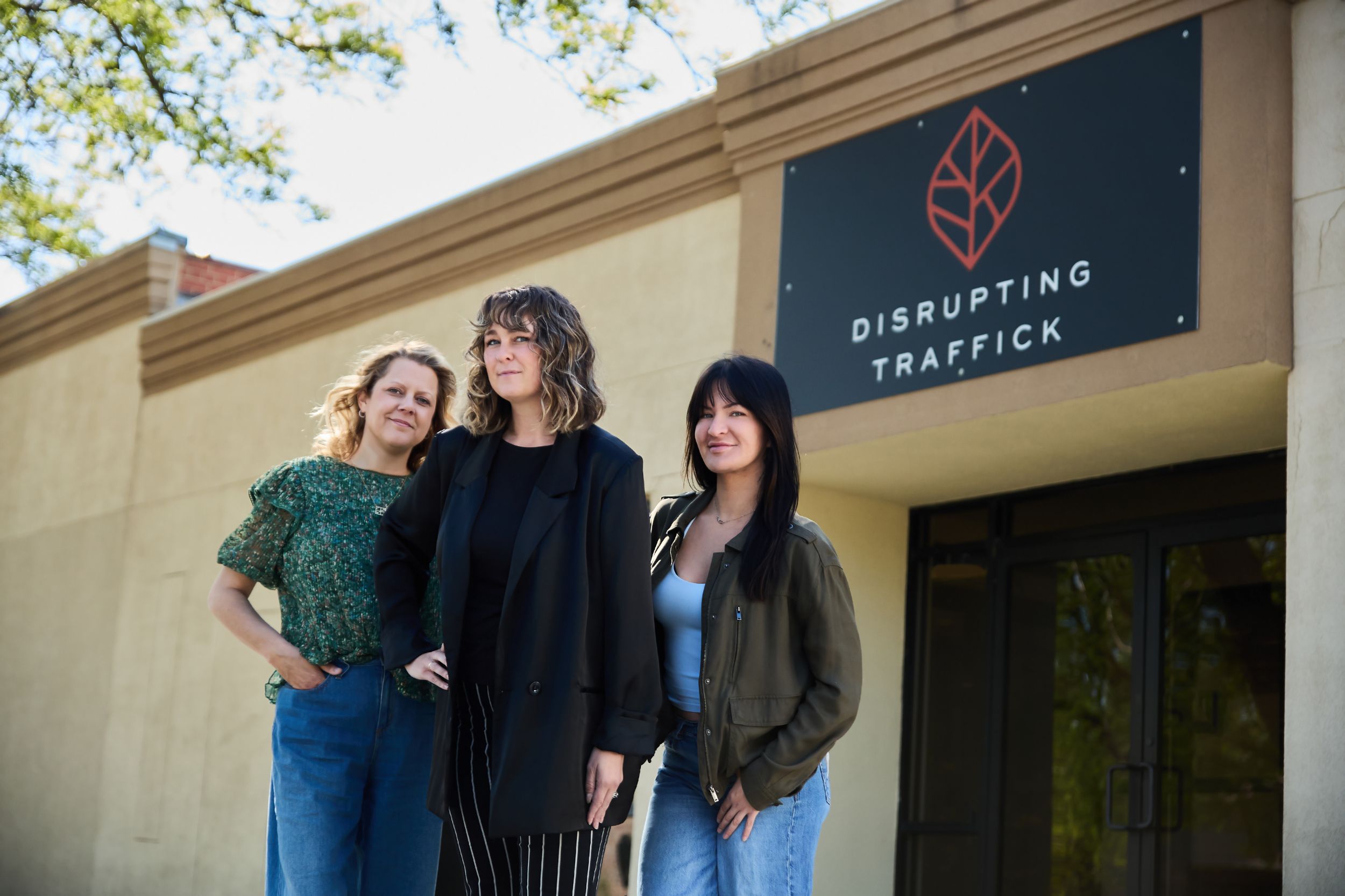 Three women stand outside the Disrupting Traffick organization office building beneath their logo sign.