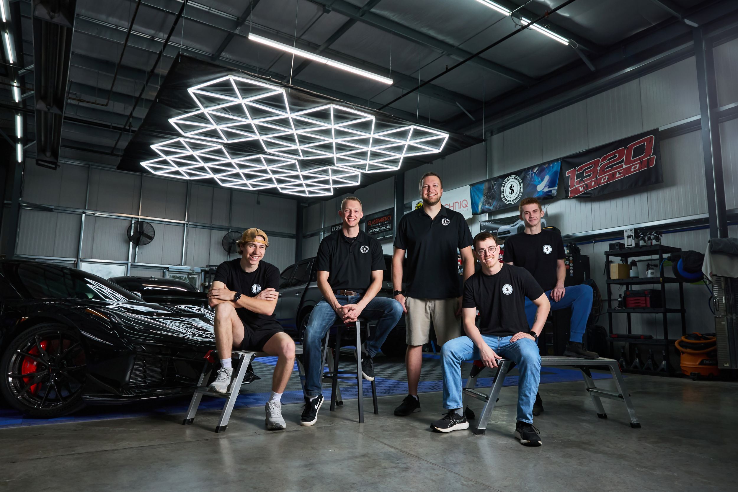 Five auto shop team members pose confidently in a modern garage with hexagonal LED lighting and a black sports car.