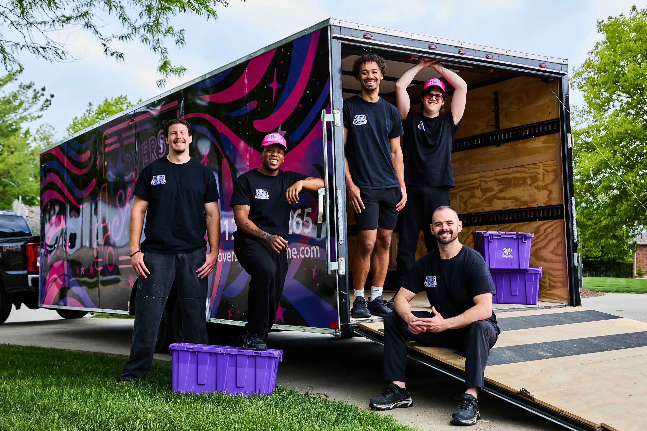 Four movers in black shirts pose proudly beside a colorful branded moving truck with purple bins on a sunny day.