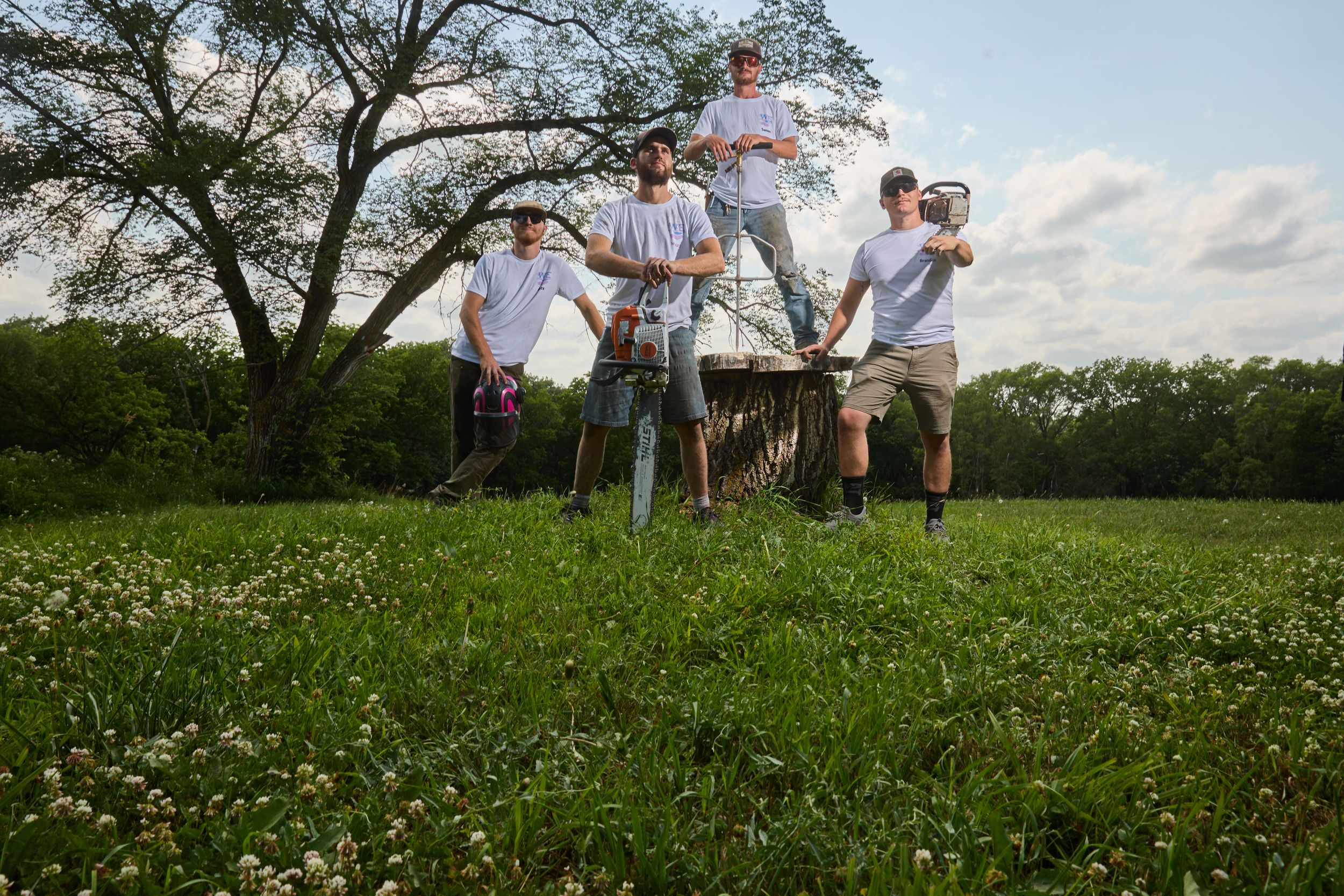 Four men posing outdoors in a green field with tools, trees, and a cloudy sky in the background.