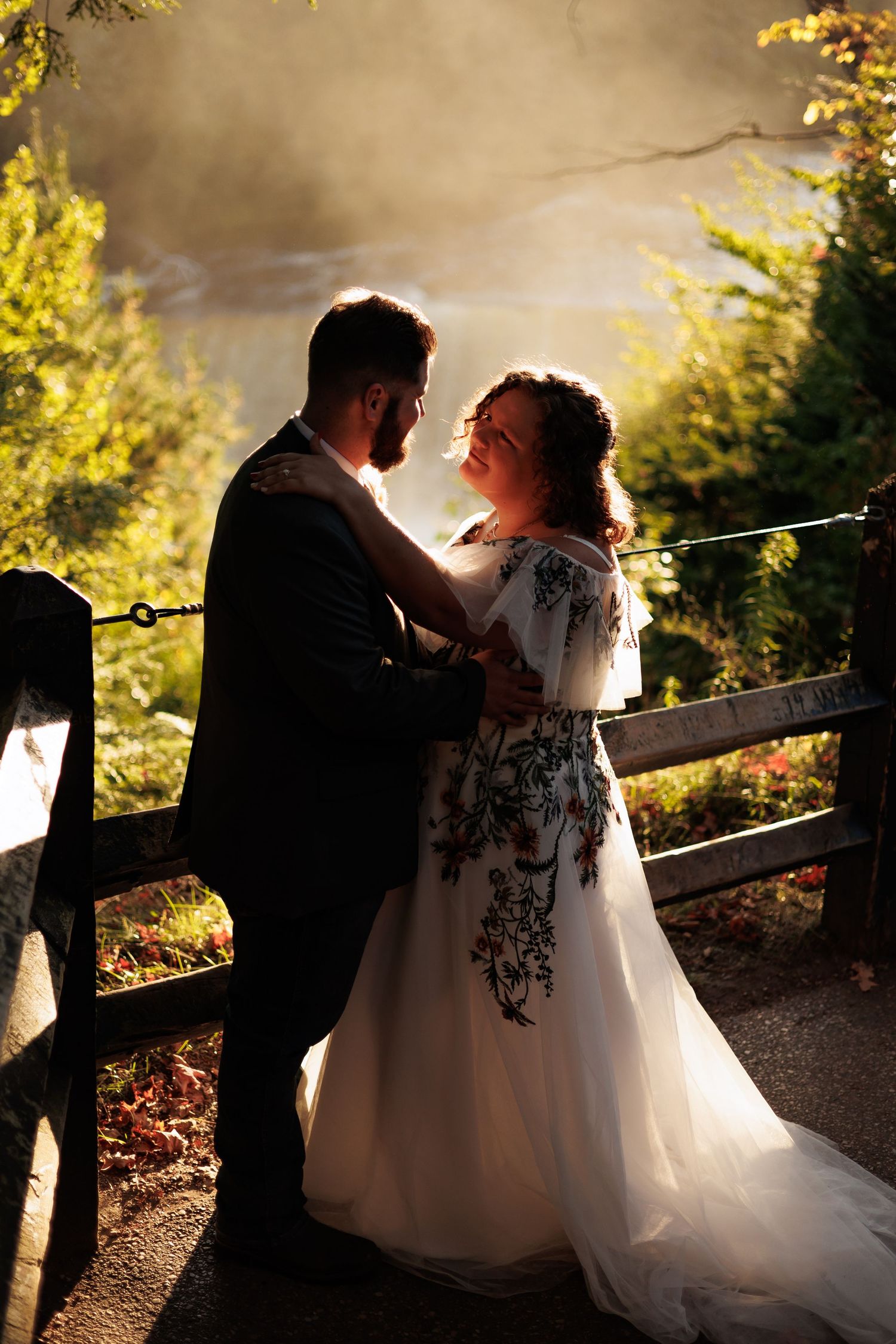Bride and groom embrace at golden hour near a wooden fence, her floral embroidered white gown glowing in the warm backlight.