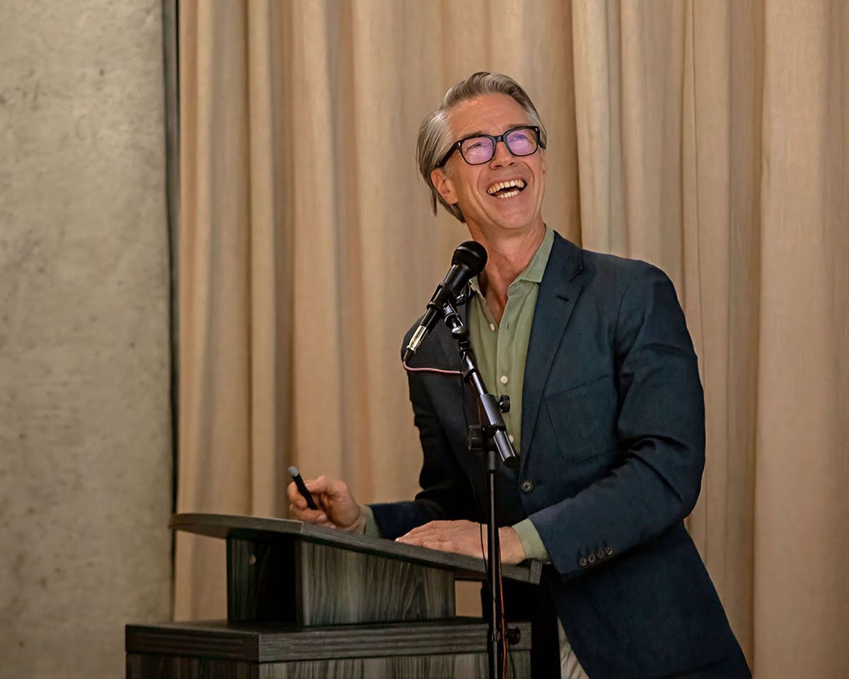 Smiling speaker at podium wearing blue blazer and pink glasses presenting at an indoor event.