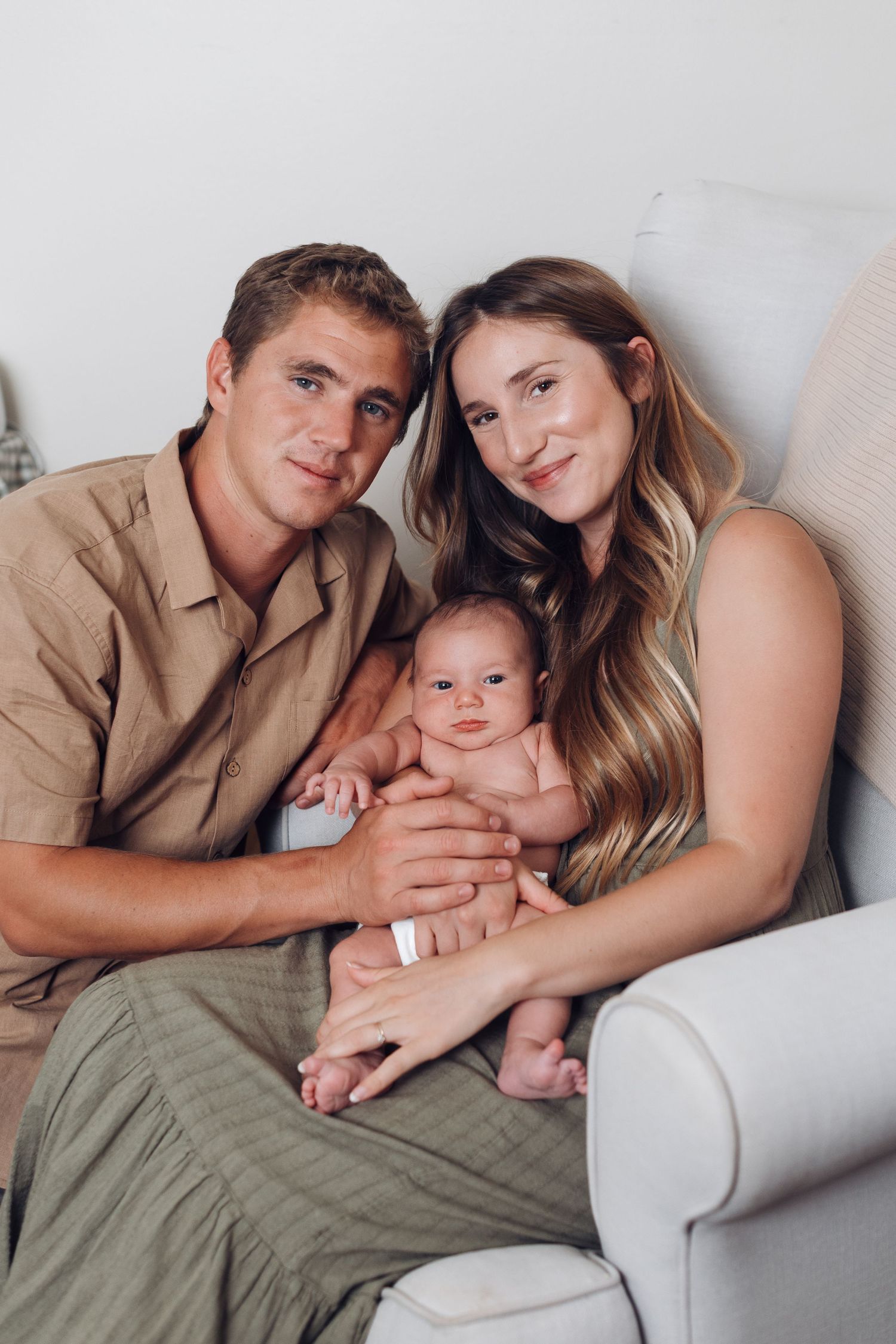 Young couple sitting in a white chair holding their newborn baby, smiling warmly in a cozy indoor setting.